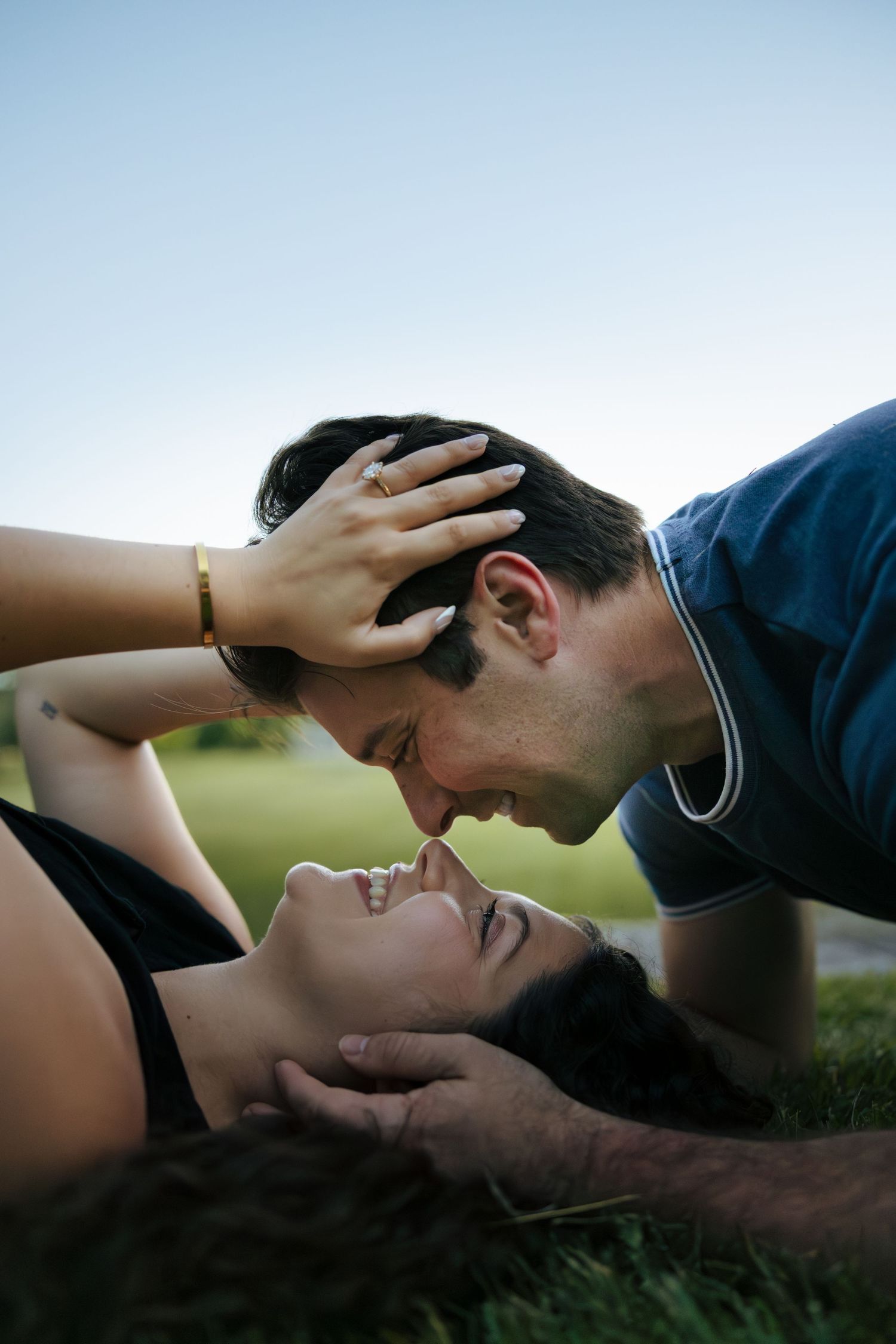 A romantic couple lies on grass, faces close together, the woman resting her hand on the mans head.