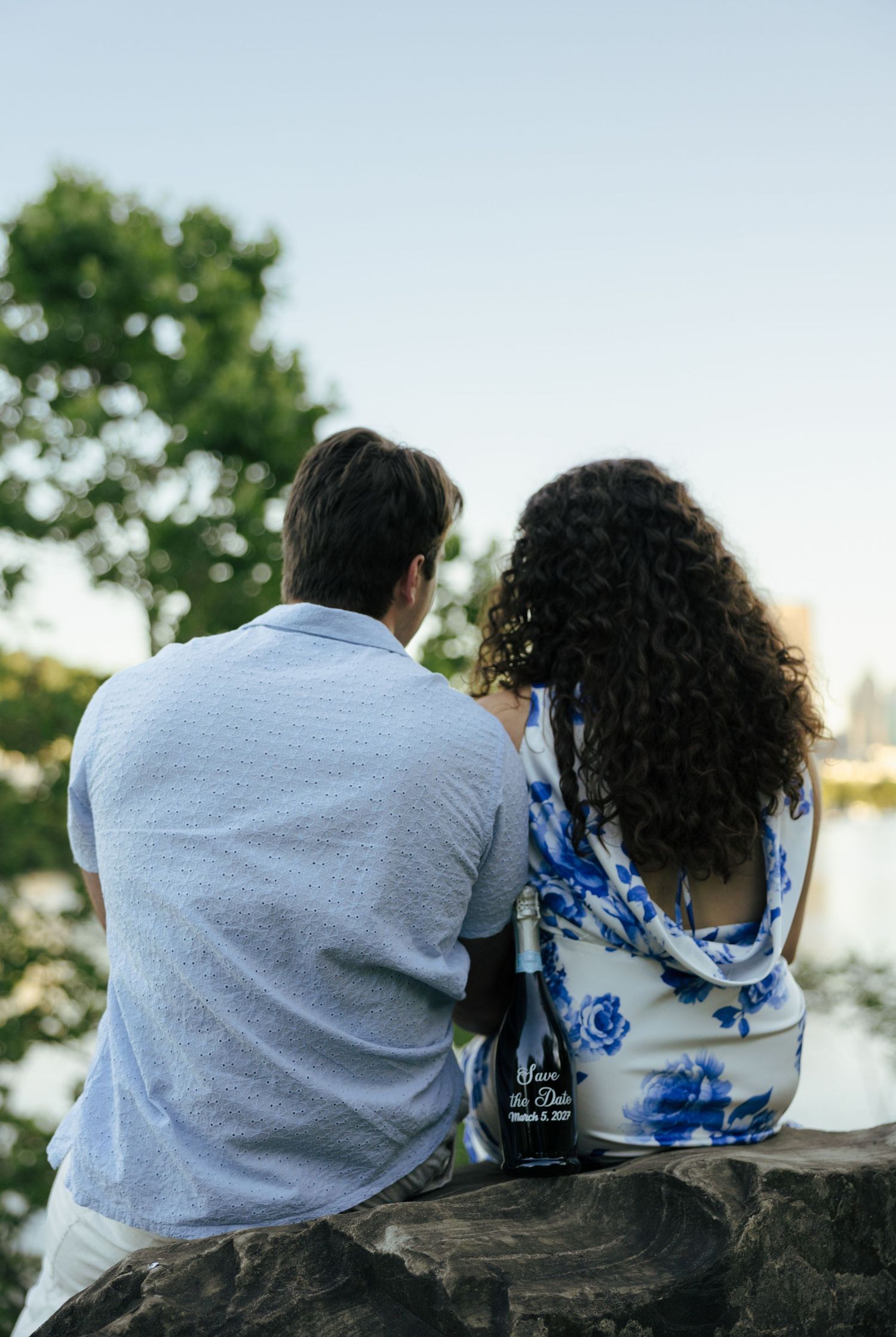 Engaged couple sitting on a stone wall from behind, holding a save the date bottle overlooking a serene lake.