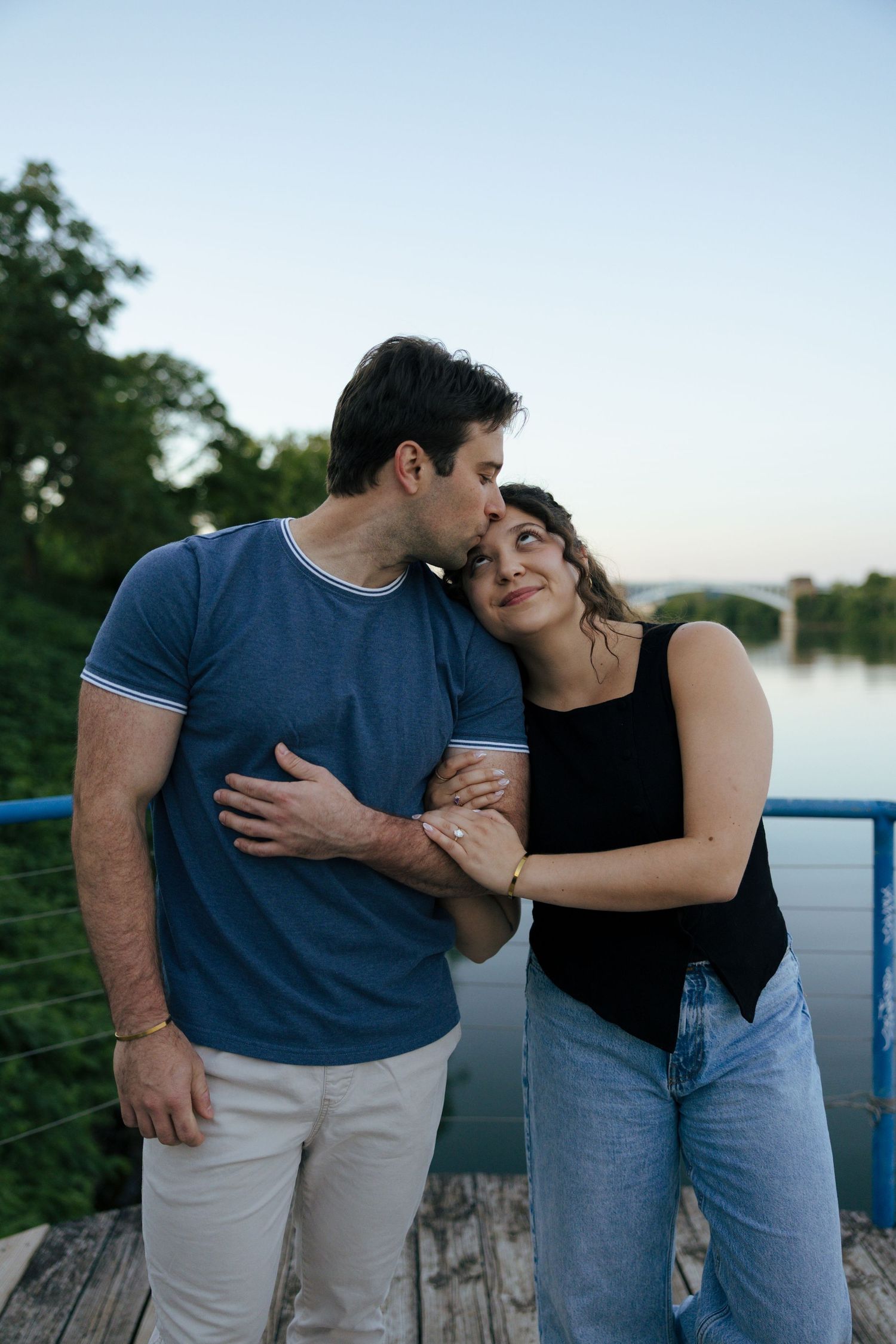 Man tenderly kissing woman on the forehead while holding hands on a scenic bridge.