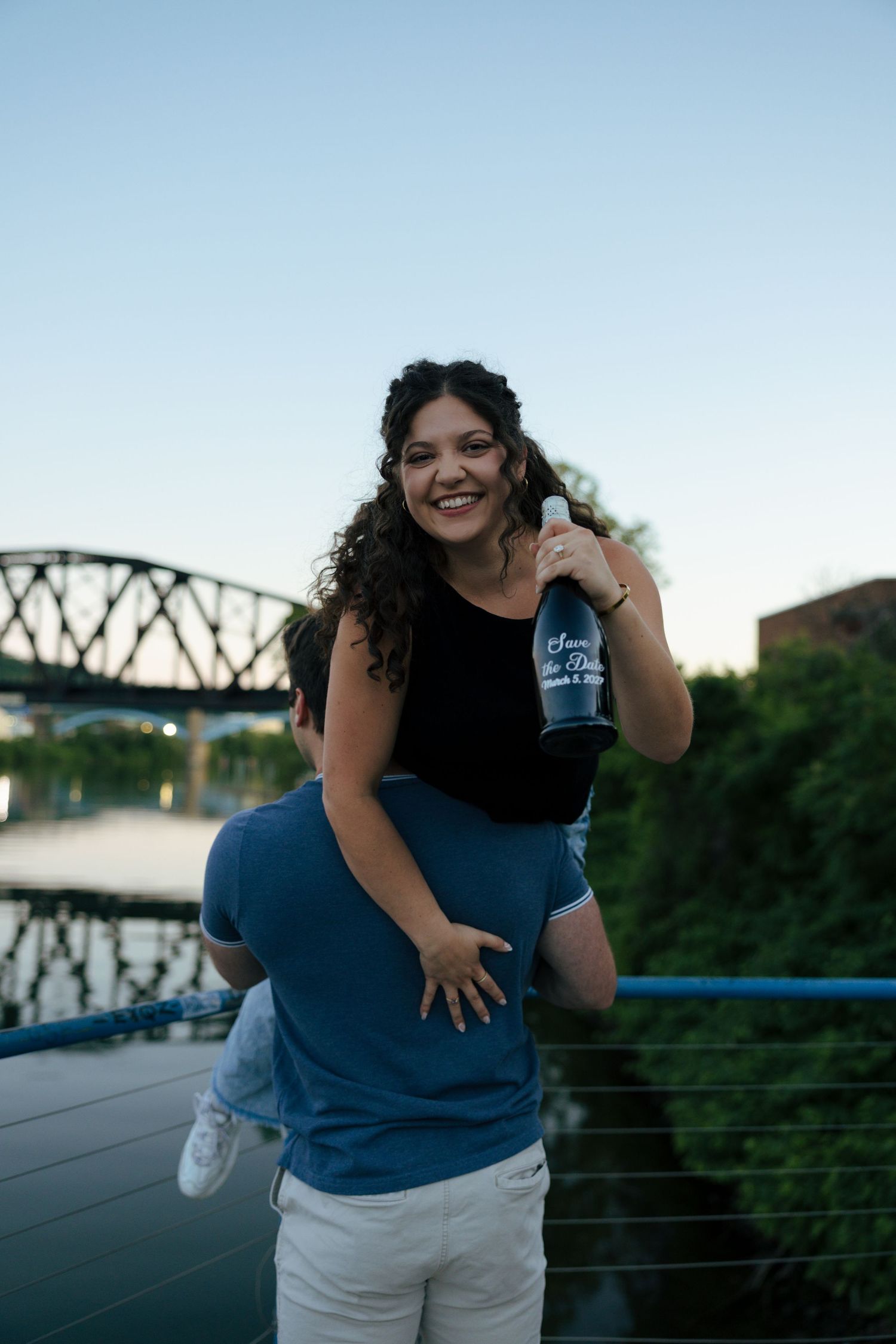 A woman smiles while getting a piggyback ride from a man on a bridge near a river at sunset.