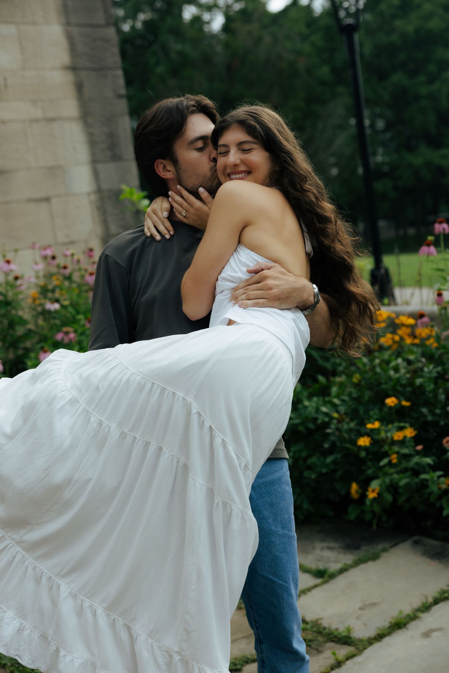A man kisses a laughing woman in a white dress while holding her in a garden.