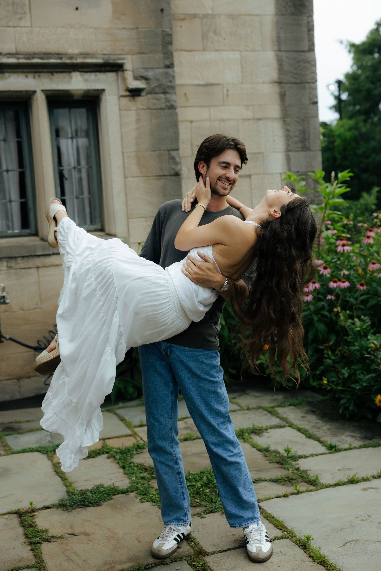 Man lifting woman in white dress outdoors near stone building, both smiling happily together.