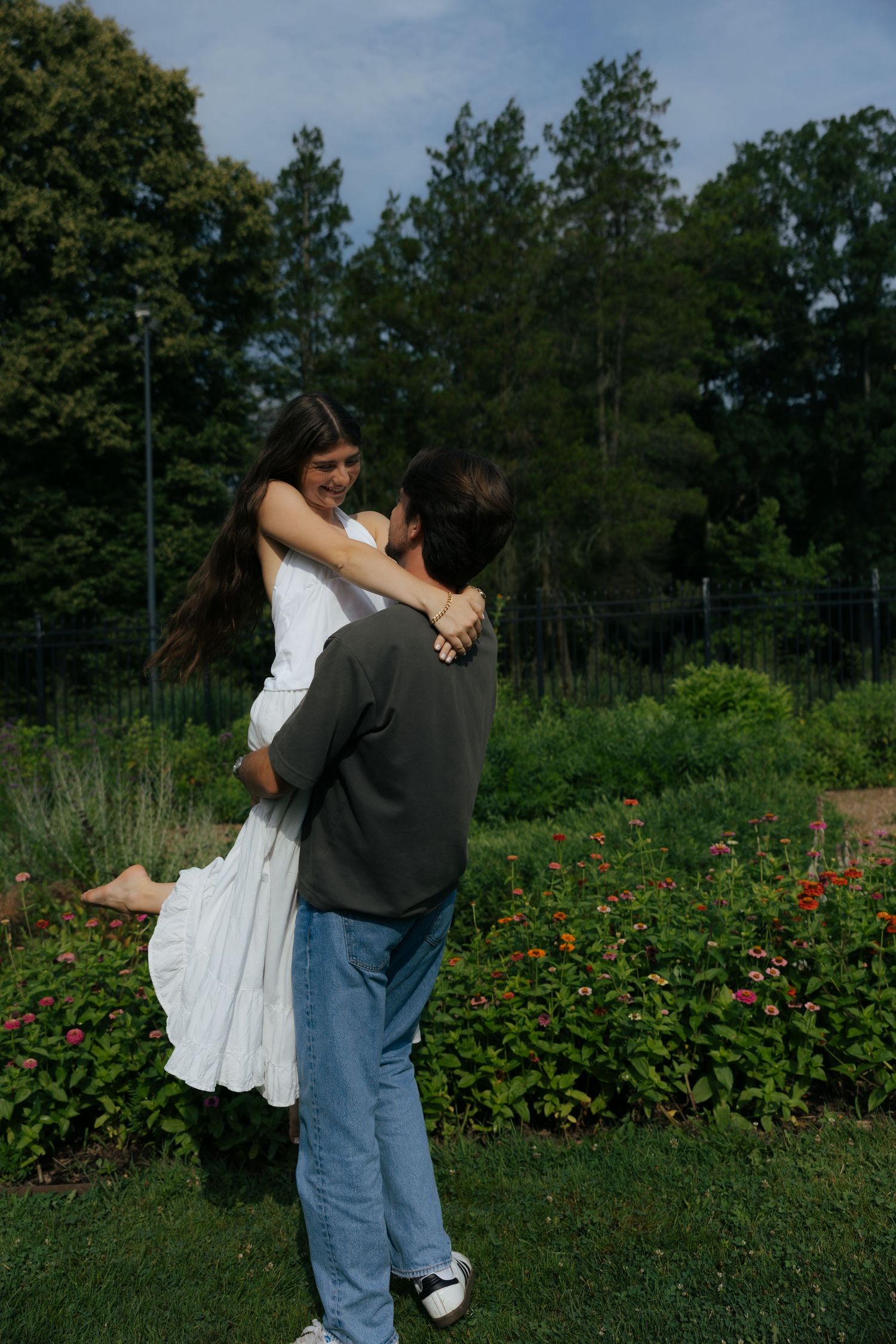 A man lifts a laughing woman in a white dress in a vibrant garden, sharing a joyful romantic moment outdoors.