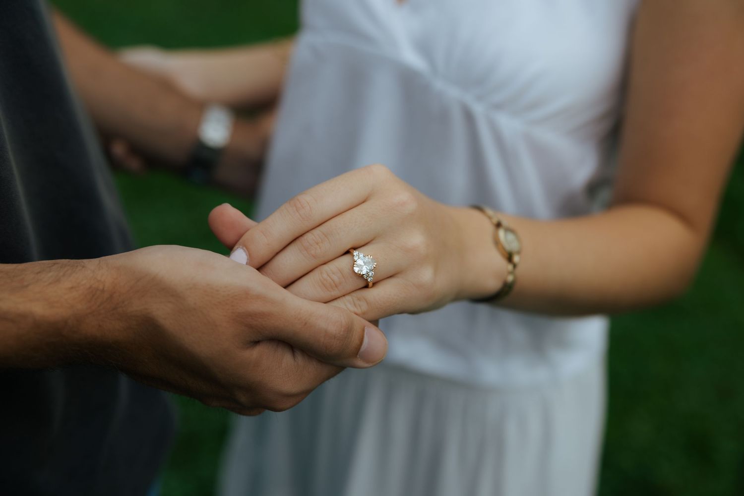 Close-up of a woman's hand wearing a diamond engagement ring, held gently by a man's hand outdoors.