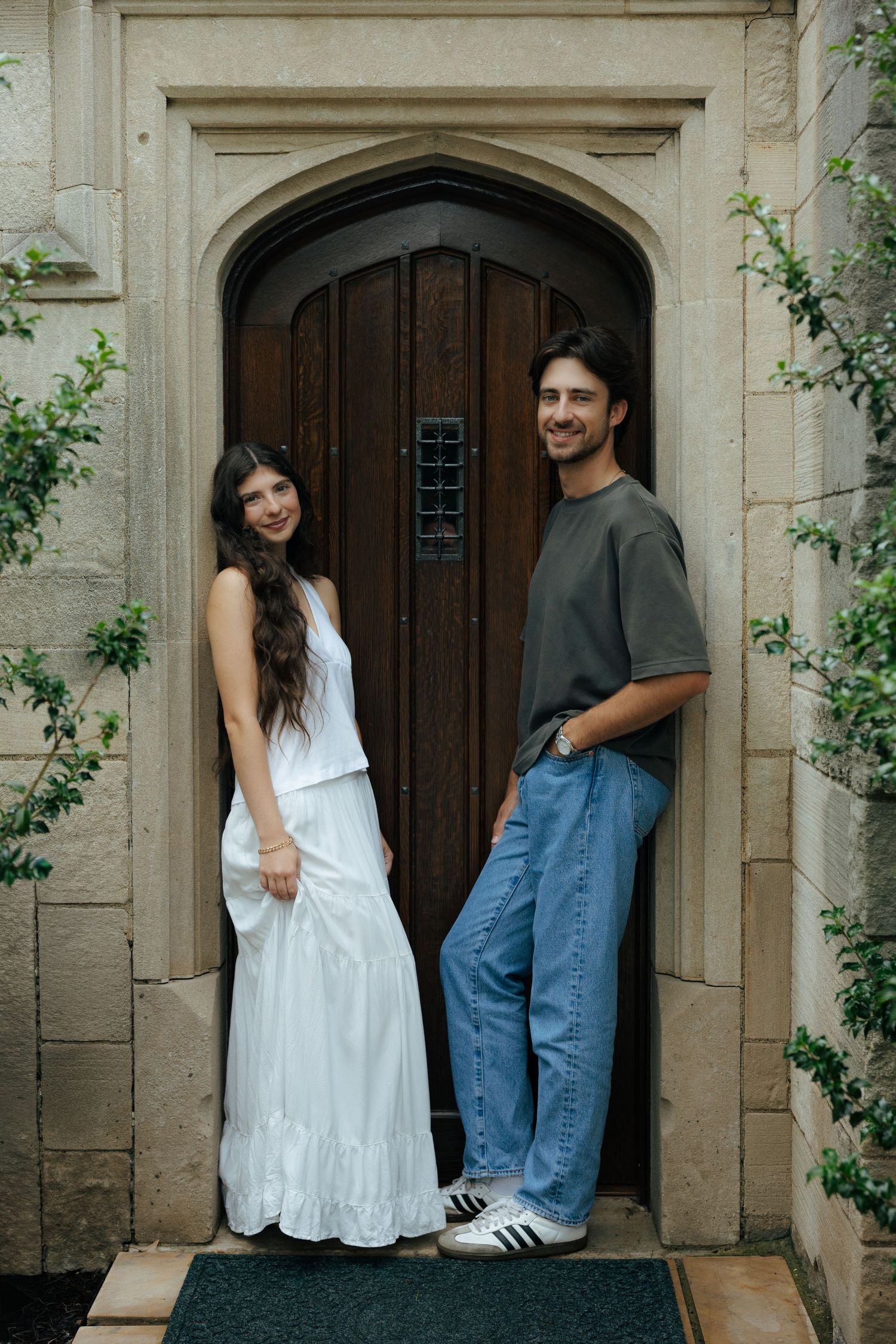 Couple posing at ornate stone arched doorway, woman in white dress, man in gray tee and jeans.