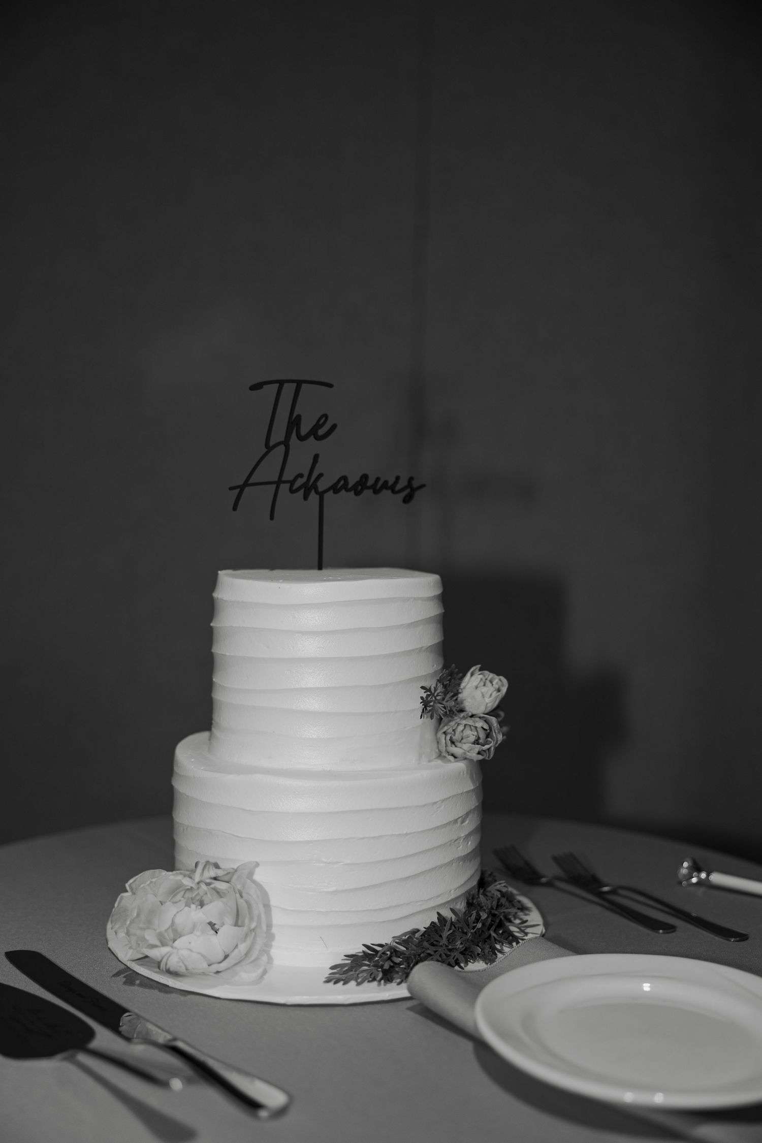 Black and white photo of a two-tier wedding cake with floral decor and The Adkasons cake topper on a table.