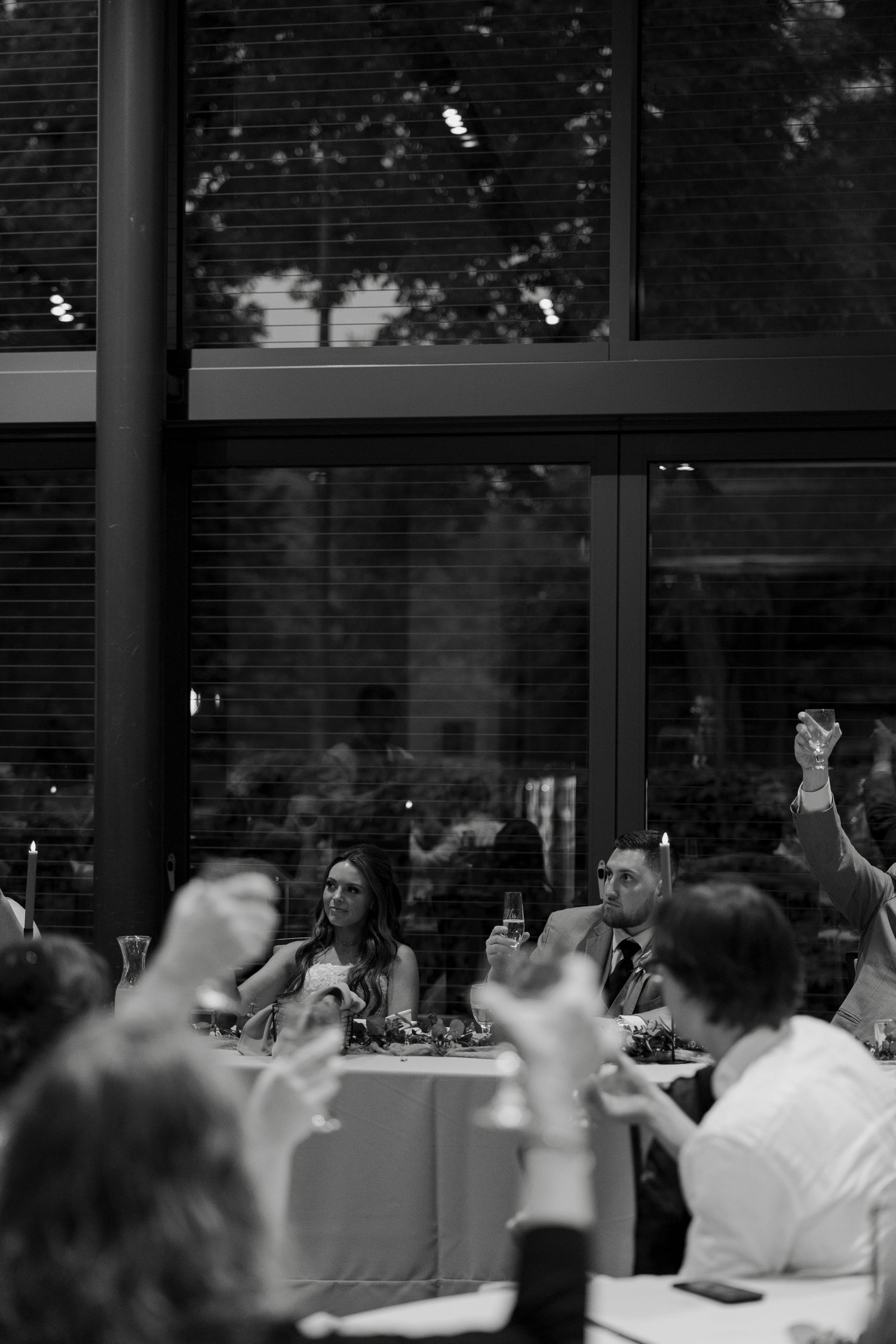 Black and white photo of wedding guests raising glasses in a toast at a reception venue with large windows.