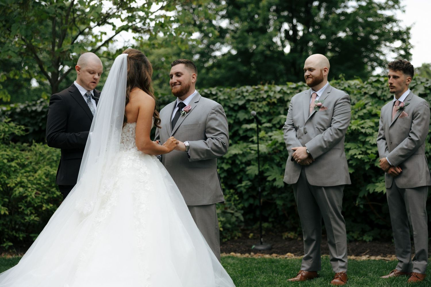 Groom holds bride's hands during outdoor wedding ceremony with groomsmen in gray suits standing nearby.
