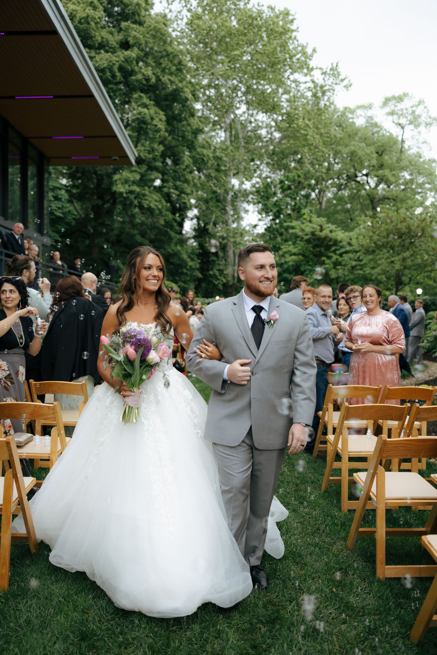 Newlyweds walk joyfully down outdoor aisle after ceremony, bride holding purple bouquet in white gown.