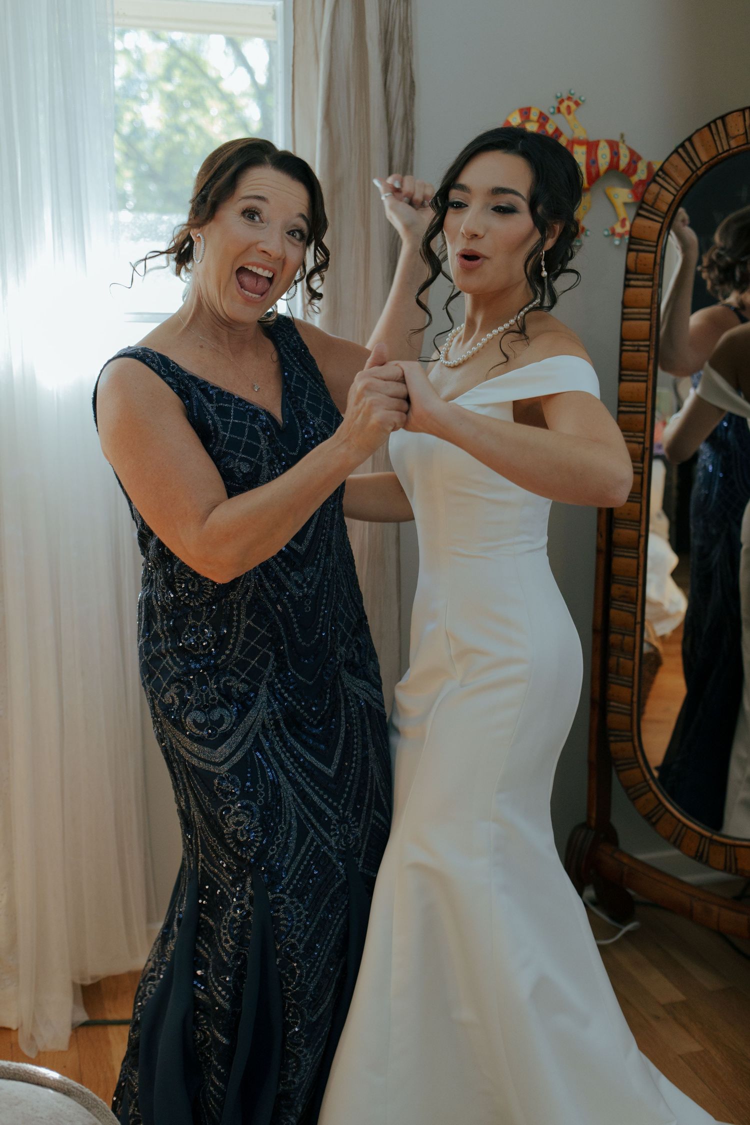 Bride in white mermaid gown laughing with woman in navy beaded dress near an arched mirror.