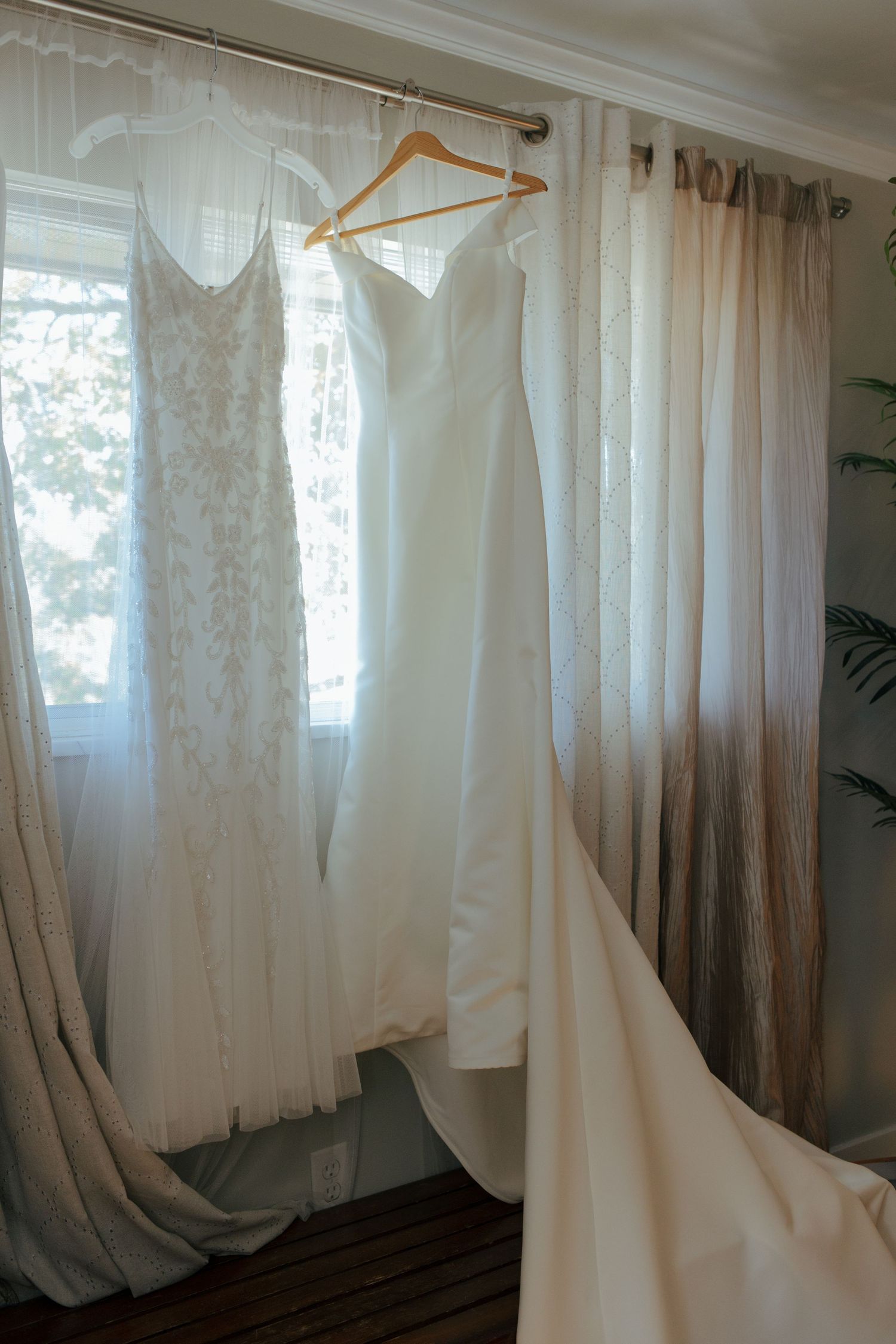 Two elegant wedding dresses hanging by a sunlit window with sheer curtains in a bridal suite.