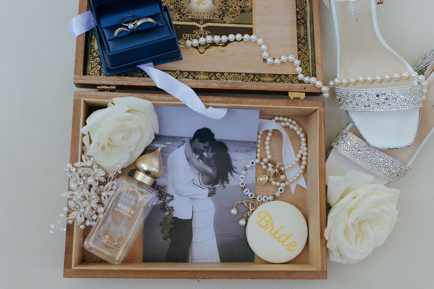Elegant bridal flat lay with wedding rings, pearls, perfume, roses, and a romantic black and white couple photo in a wooden box.