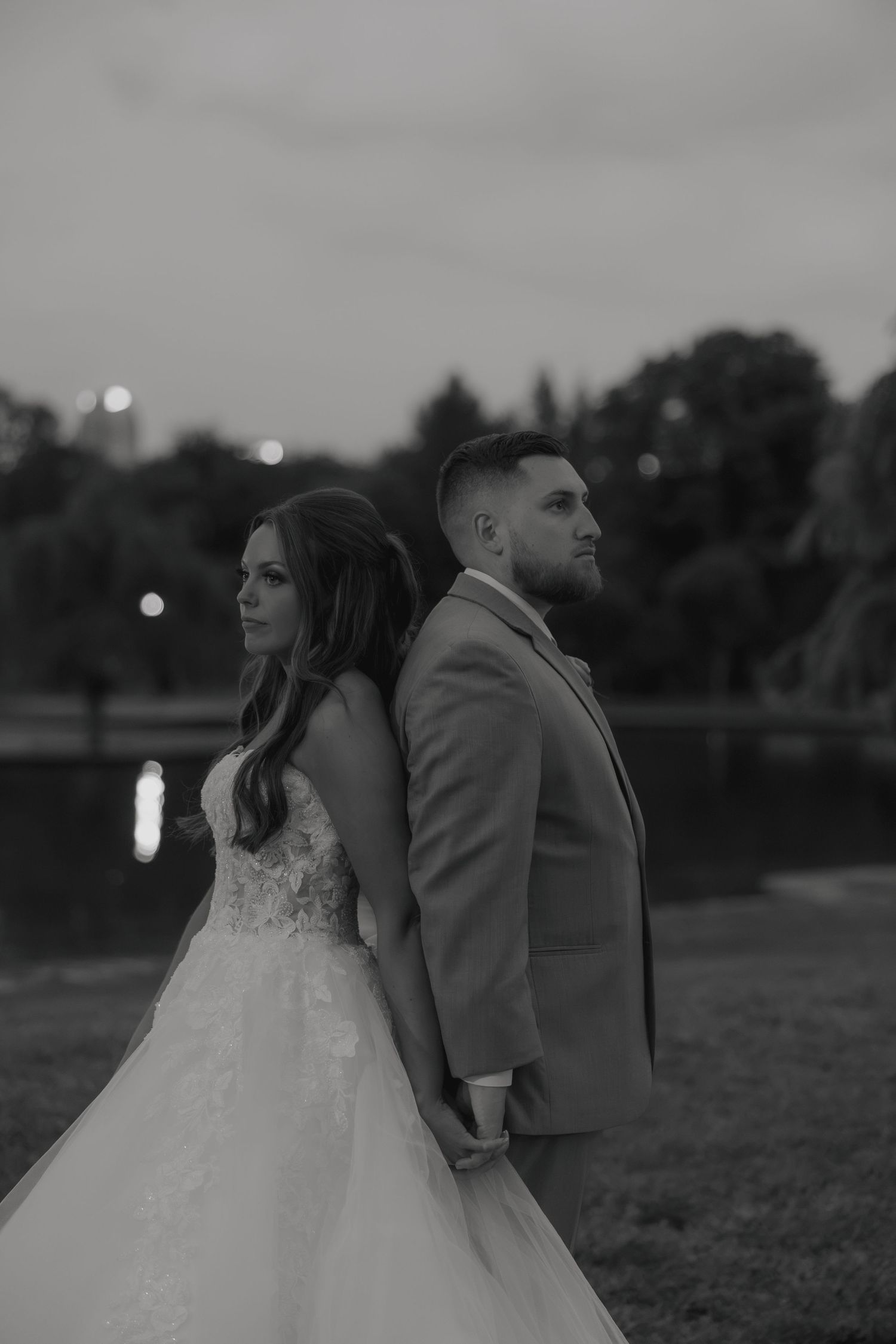 Black and white wedding photo of bride and groom standing back to back at dusk near a lake.