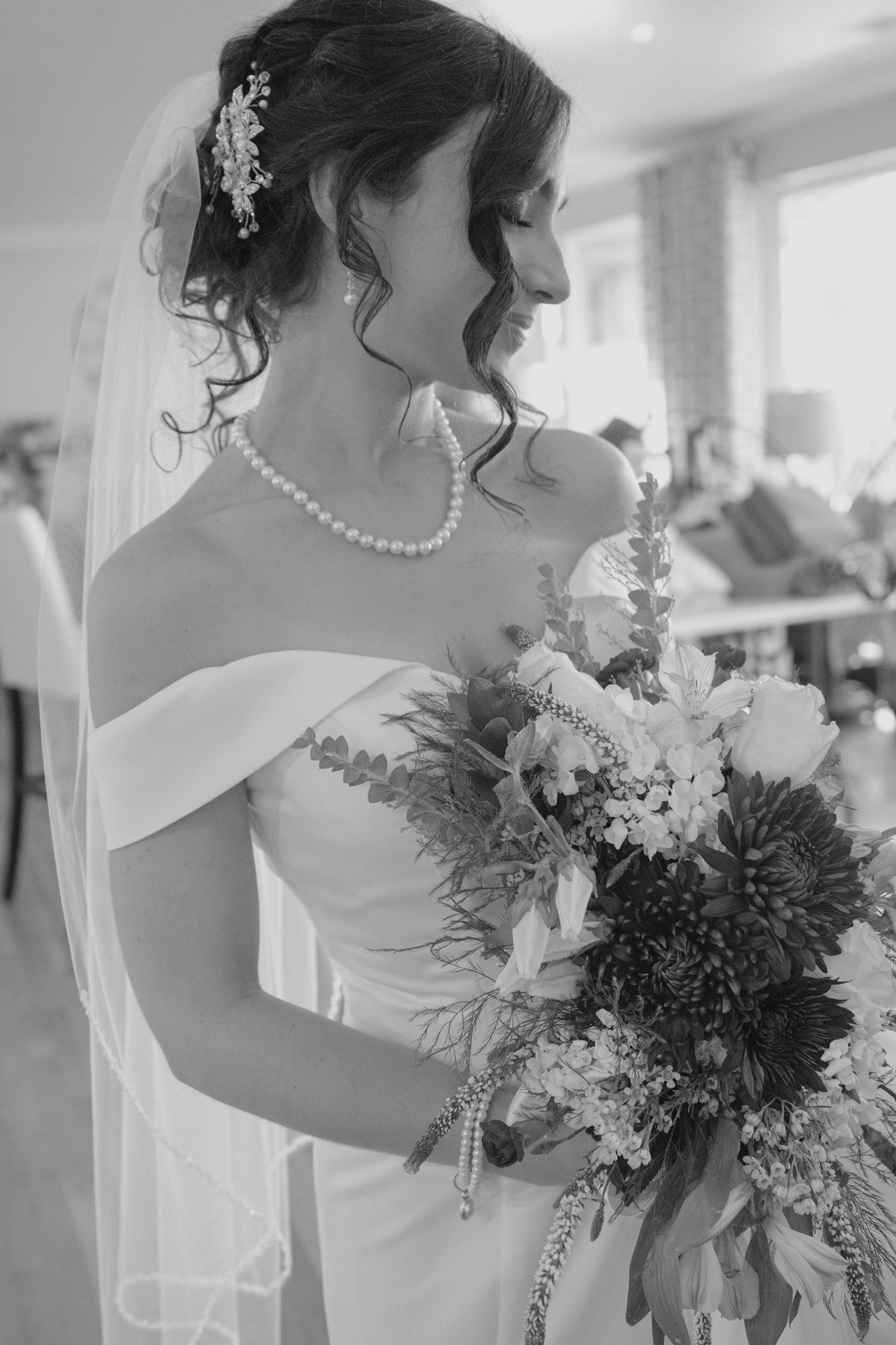 Black and white photo of a bride in off-shoulder gown, pearl necklace, veil, and floral bouquet, smiling softly indoors.