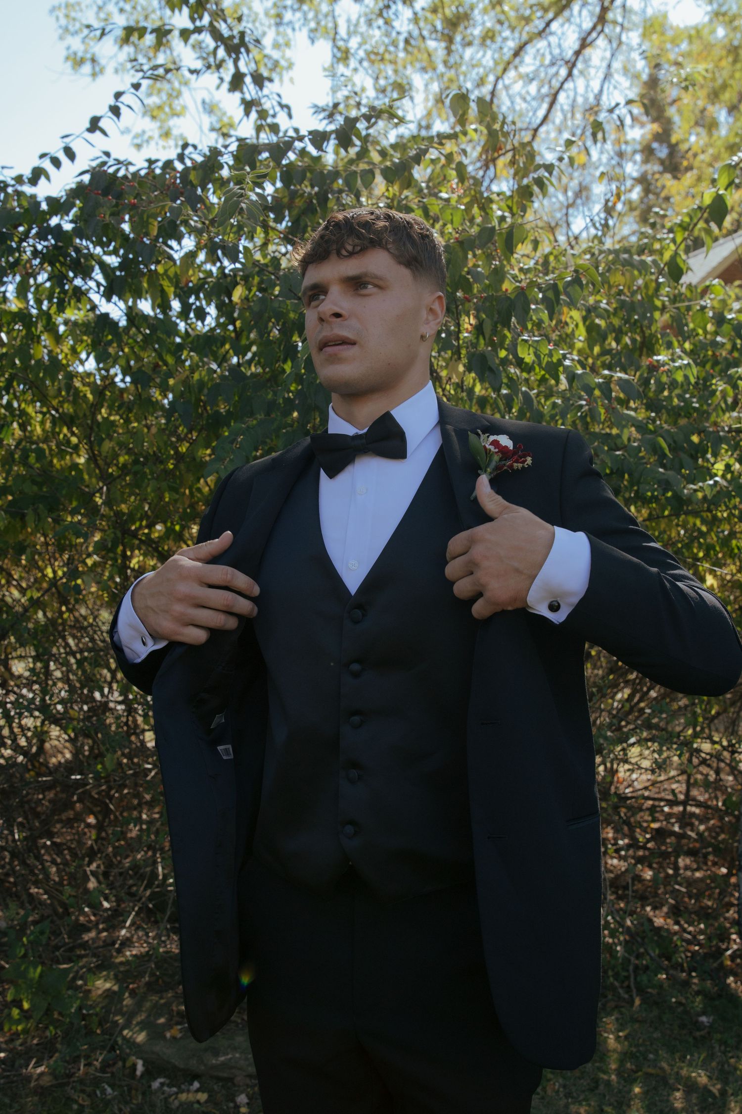 Dapper groom in black tuxedo with bow tie and boutonniere adjusts jacket lapels outdoors among lush greenery.