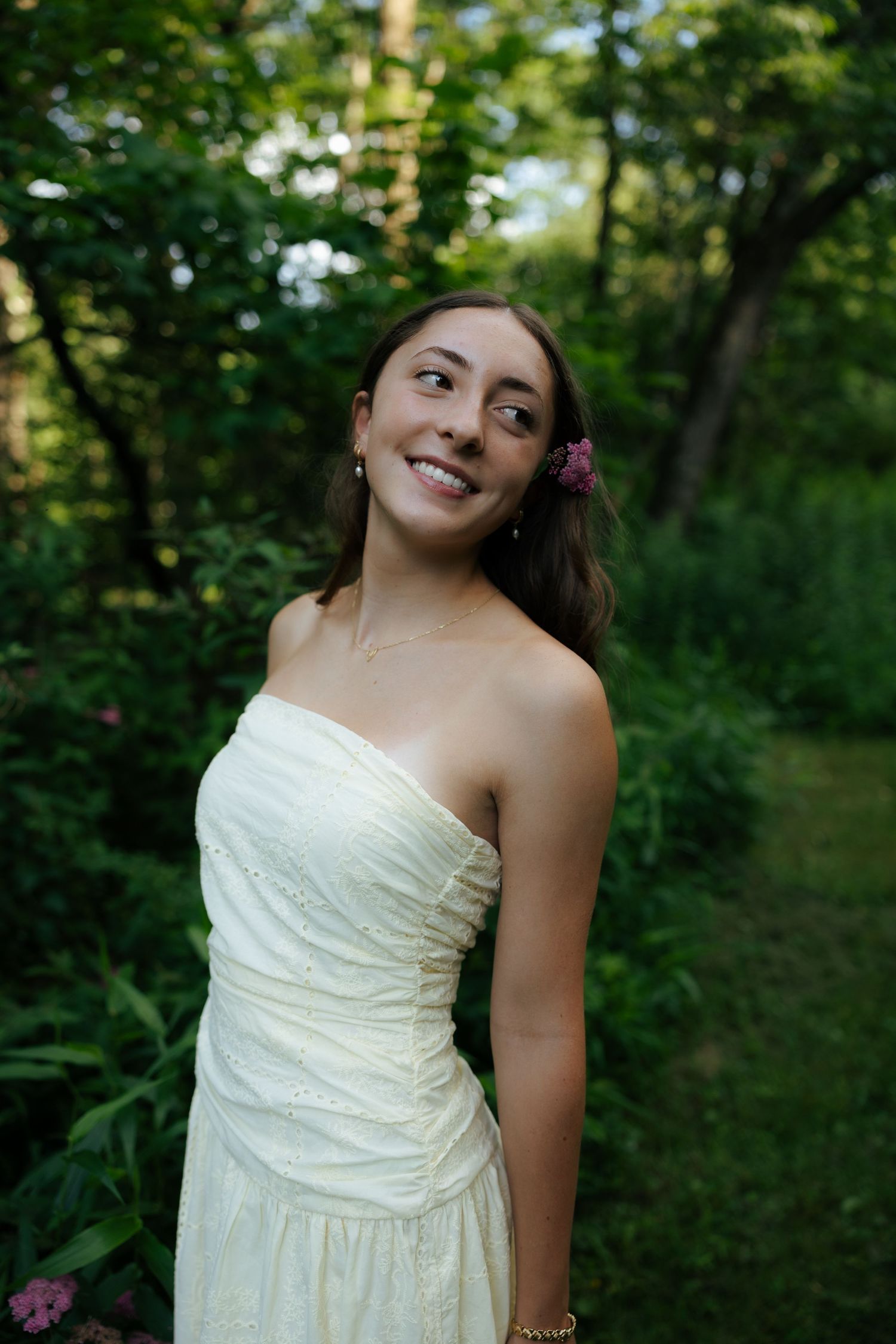 Smiling young woman in strapless white dress with flower in hair standing in green garden.