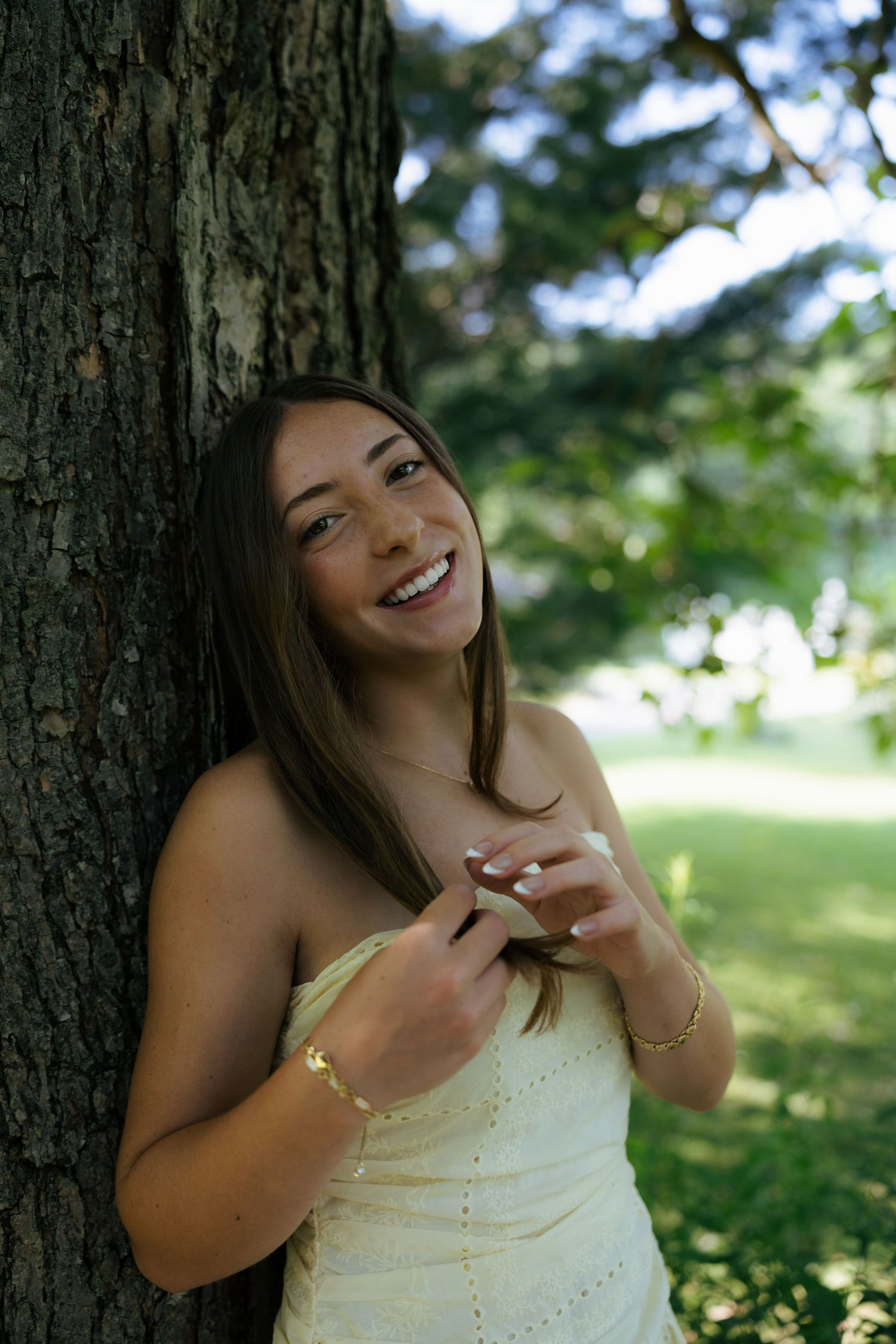 Young woman in white strapless dress smiling against tree trunk in park.