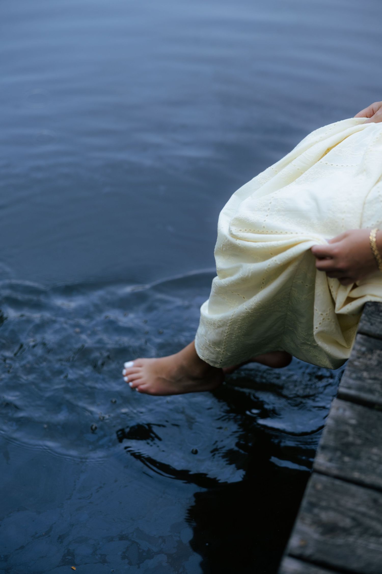 Close-up of barefoot woman in cream dress dipping feet into calm lake water from wooden dock.