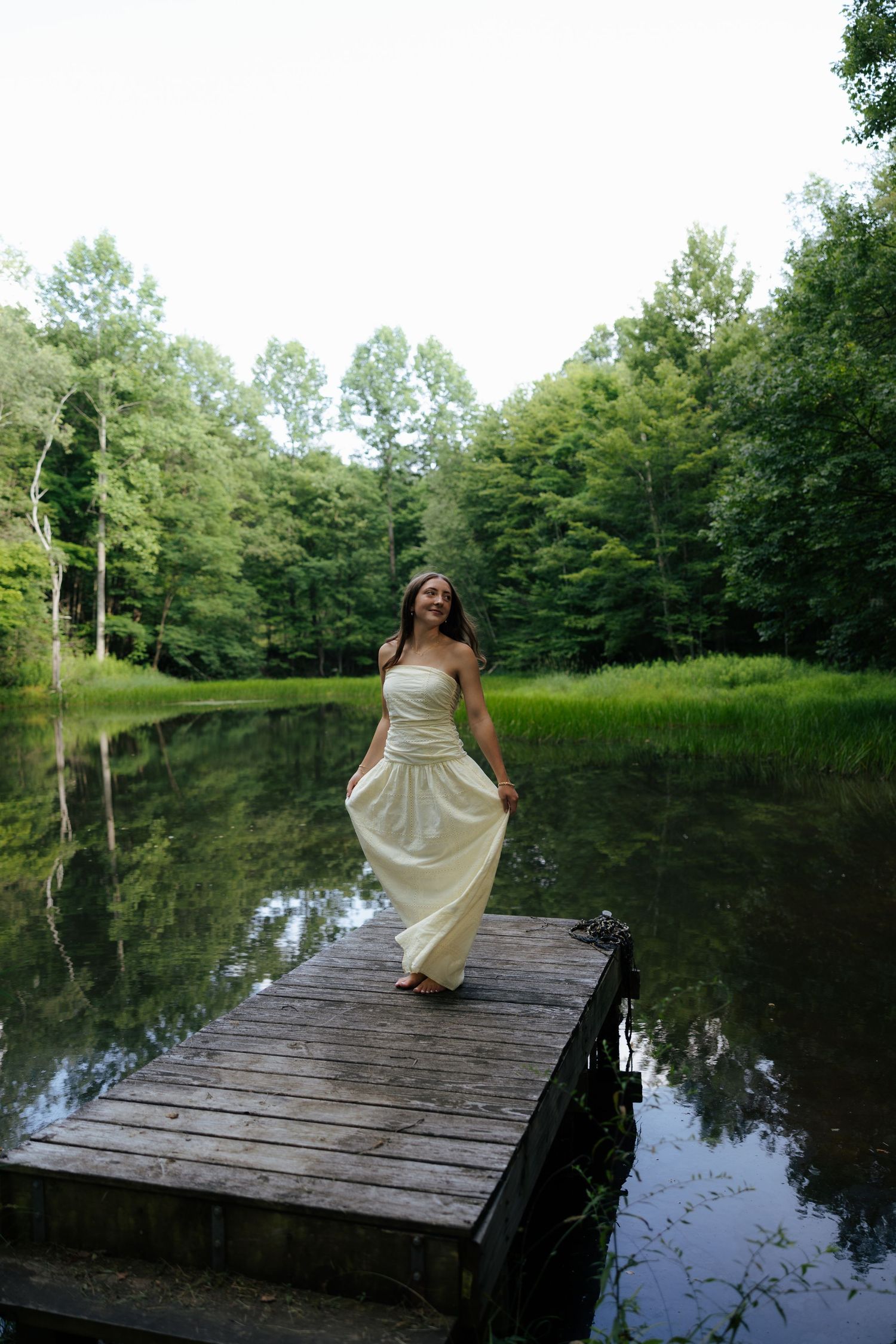 Young woman in white dress standing on wooden dock by calm reflective lake in forest.