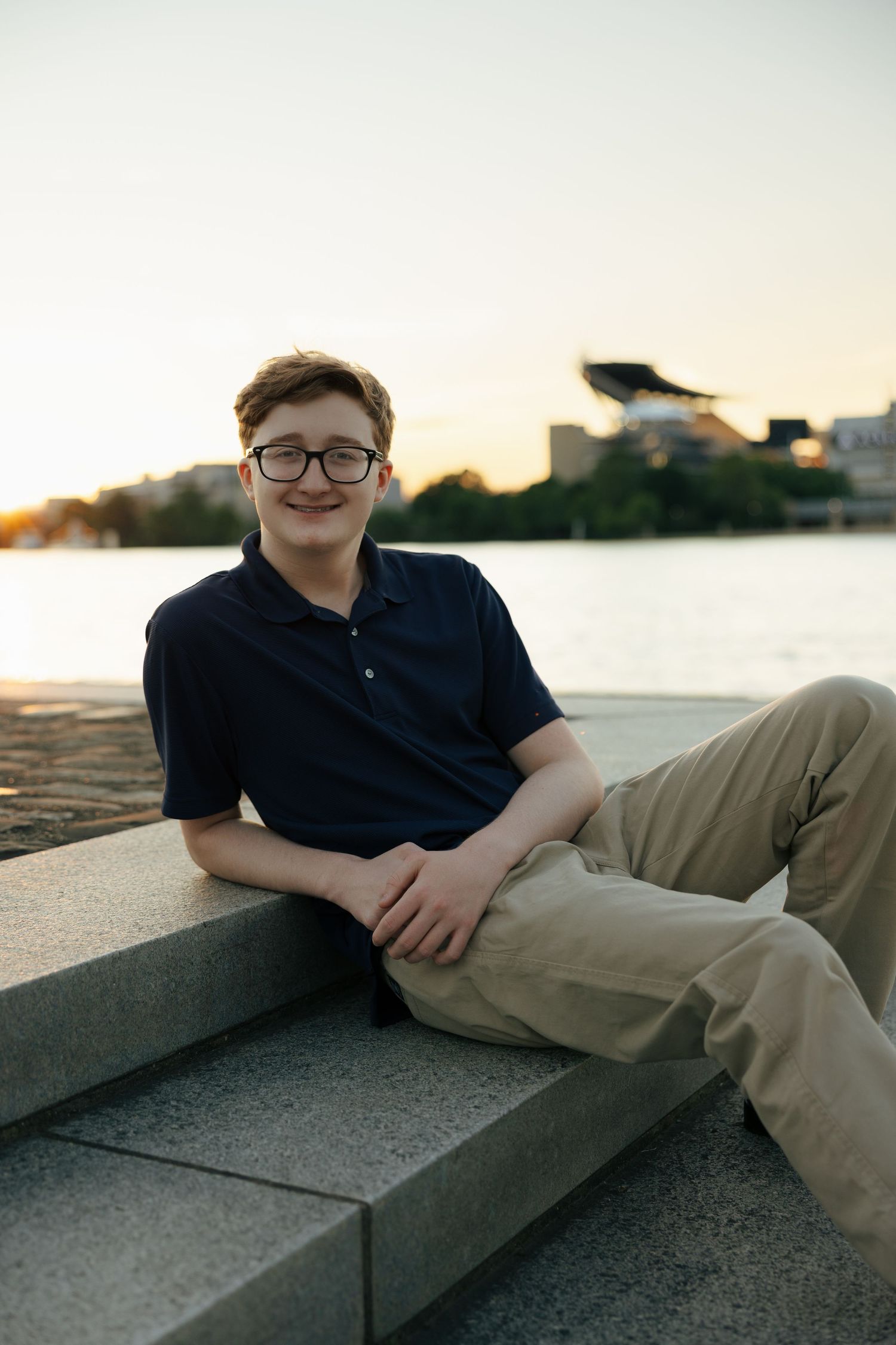 Teen boy in glasses and navy polo reclining on granite steps by river during golden hour sunset.