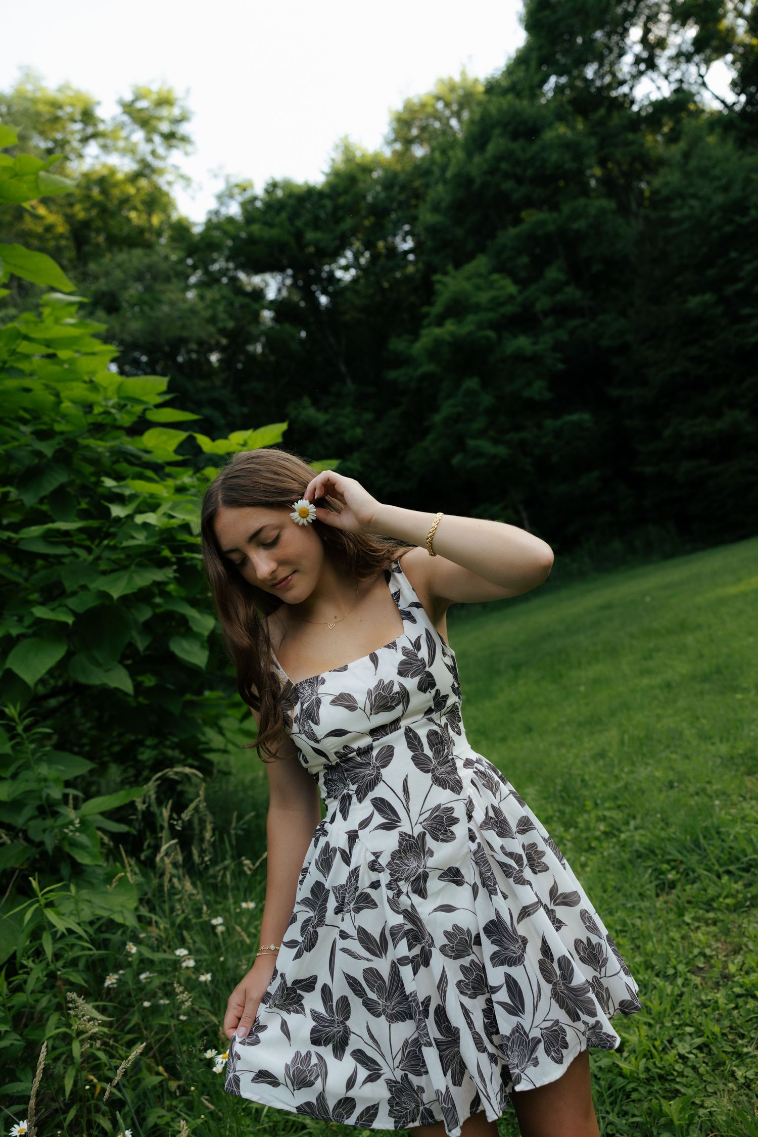 A young woman in a floral sundress stands near green bushes, adjusting her hair in a park.