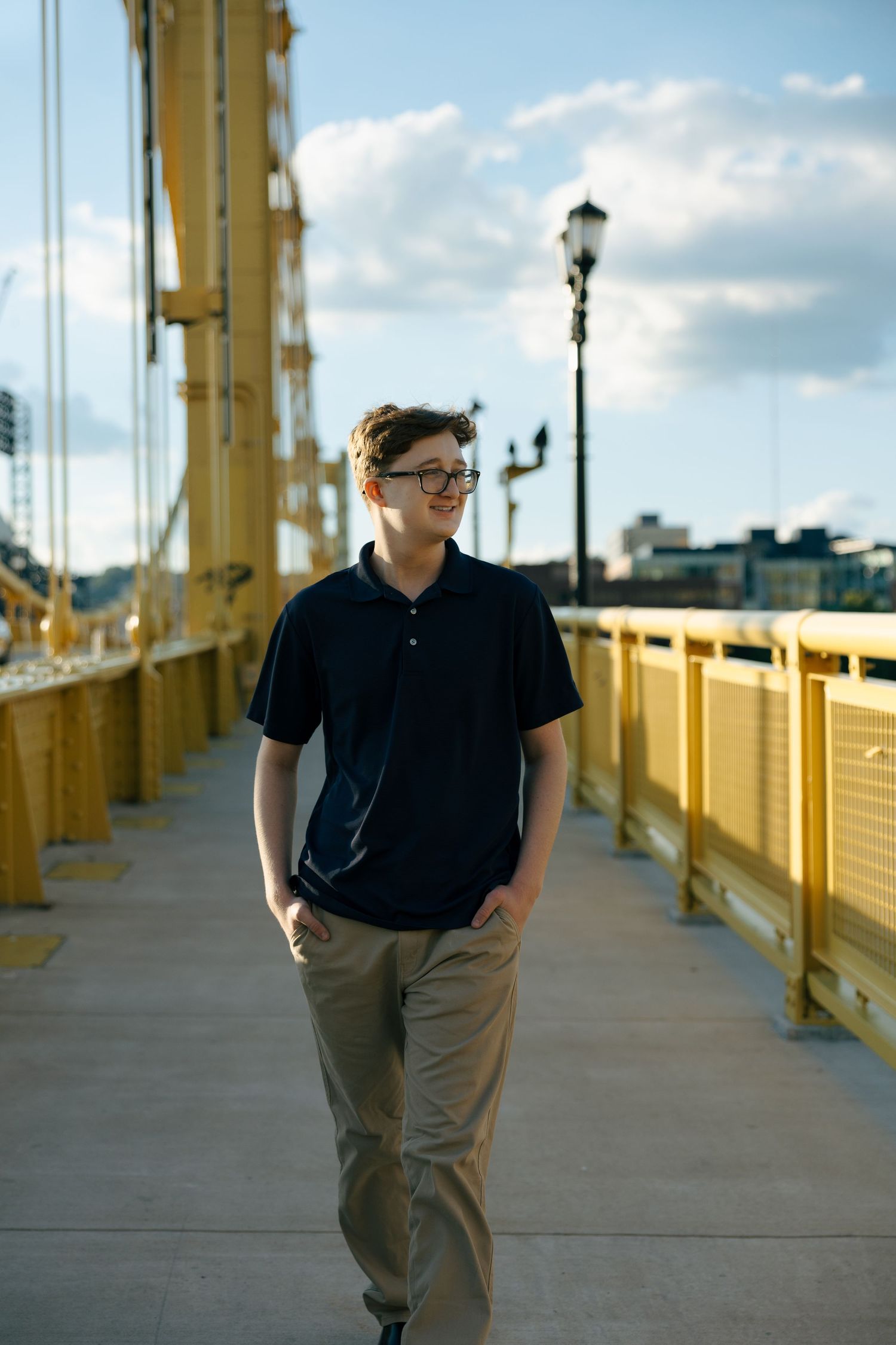 Young man in black polo and khaki pants walks on a yellow bridge, smiling, wearing glasses on a sunny day.