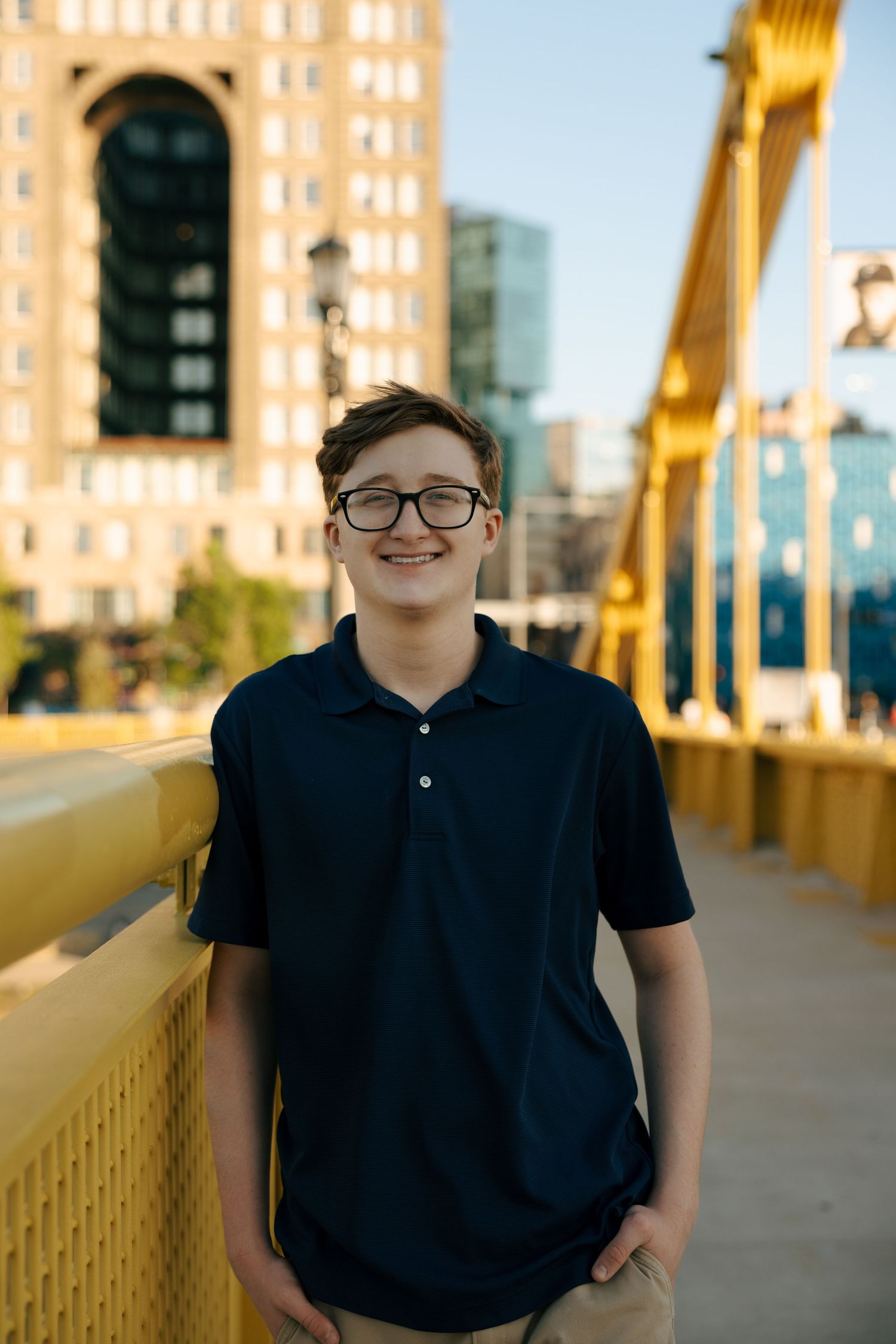 Smiling teenage boy wearing glasses and navy polo shirt standing on a yellow bridge with city buildings behind him.