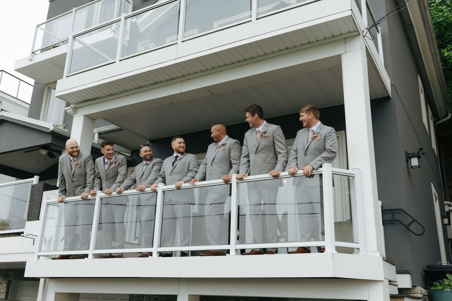 Groomsmen in matching gray suits standing on a white balcony of a modern building during a wedding celebration.