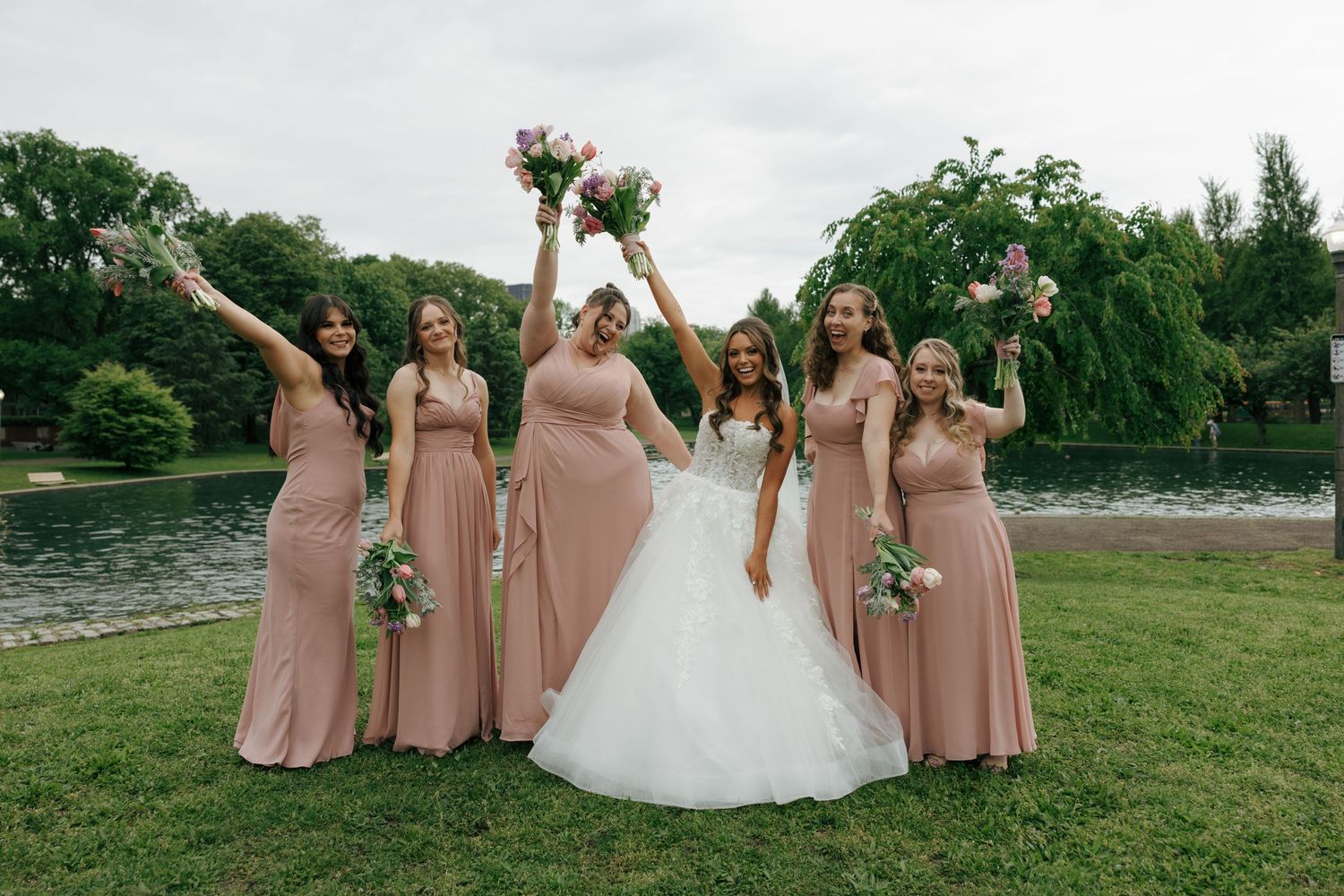 Bride in white gown with five bridesmaids in dusty rose dresses celebrating outdoors near a pond.