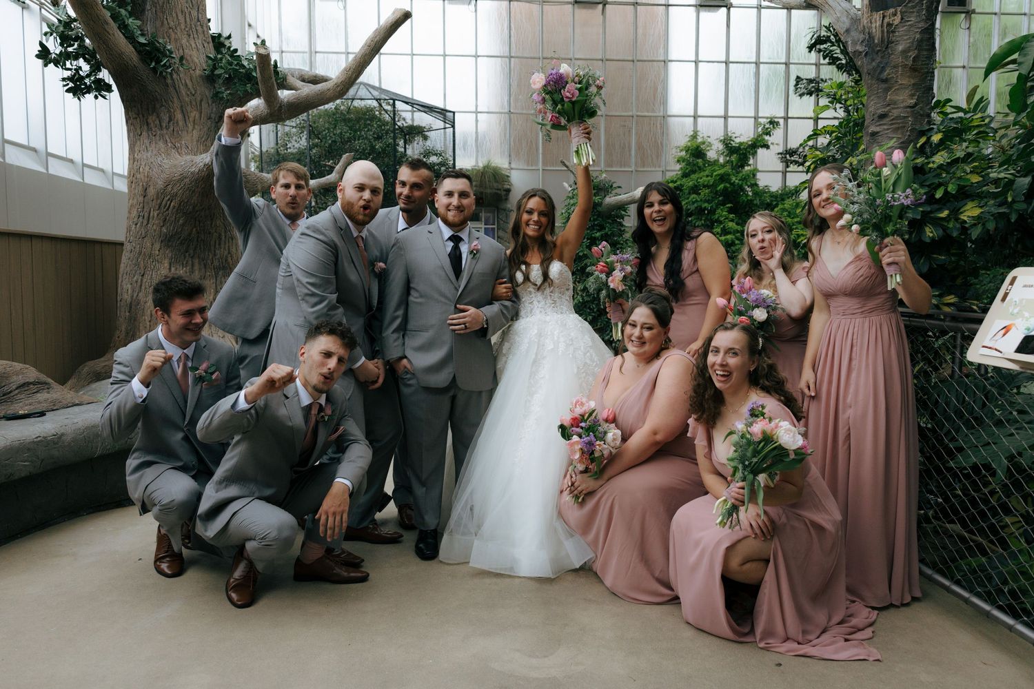 Wedding party poses in greenhouse venue, groomsmen in gray suits, bridesmaids in dusty rose gowns, bride in white gown holding bouquet.