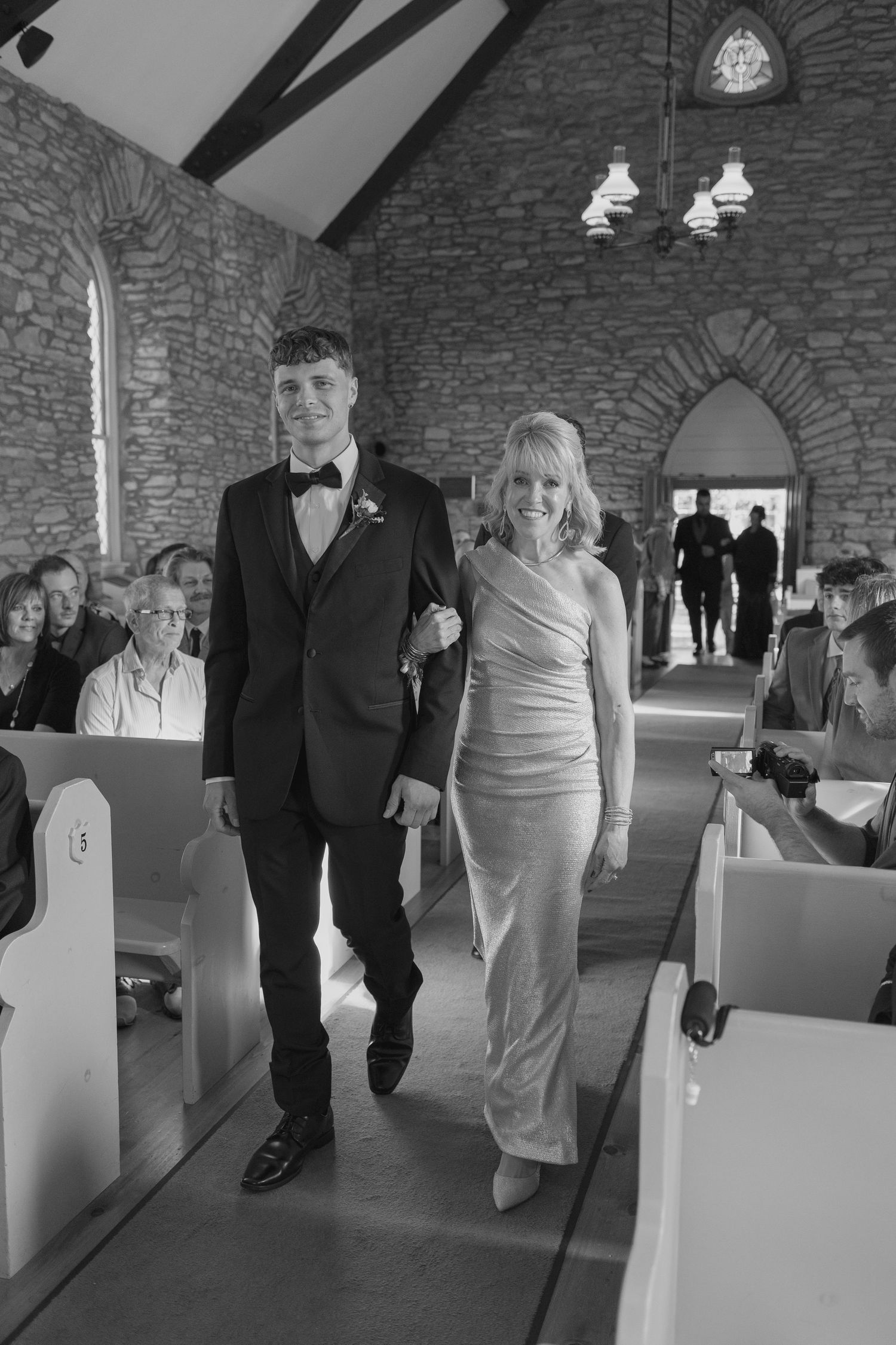 Groom in black tuxedo escorts elegantly dressed woman down church aisle during wedding ceremony.
