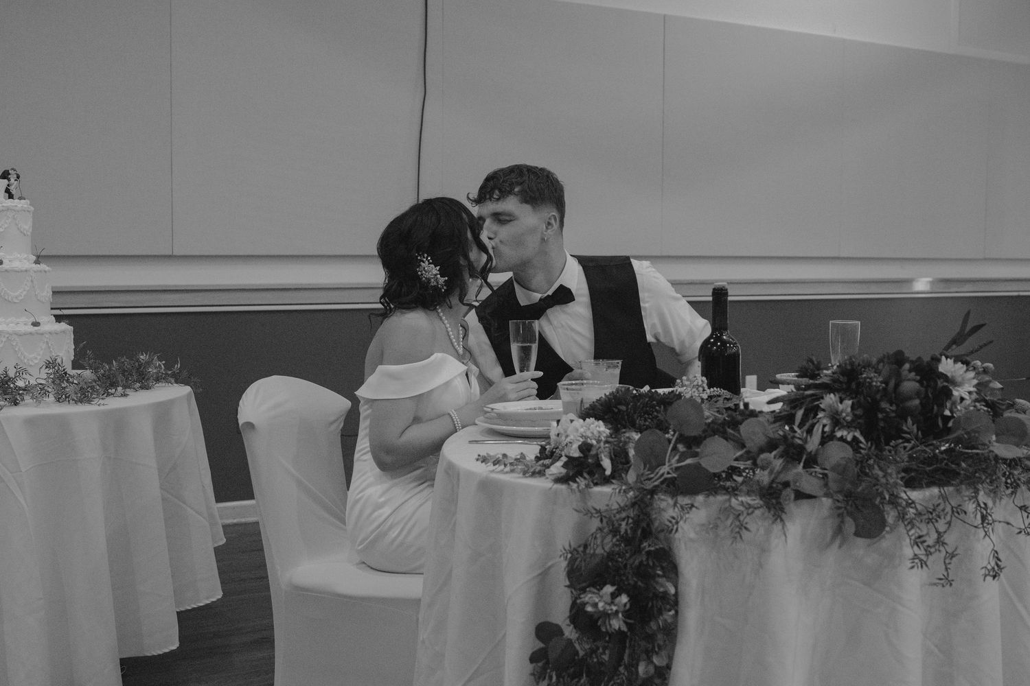 Bride and groom sharing a kiss at sweetheart table with wedding cake nearby in black and white.