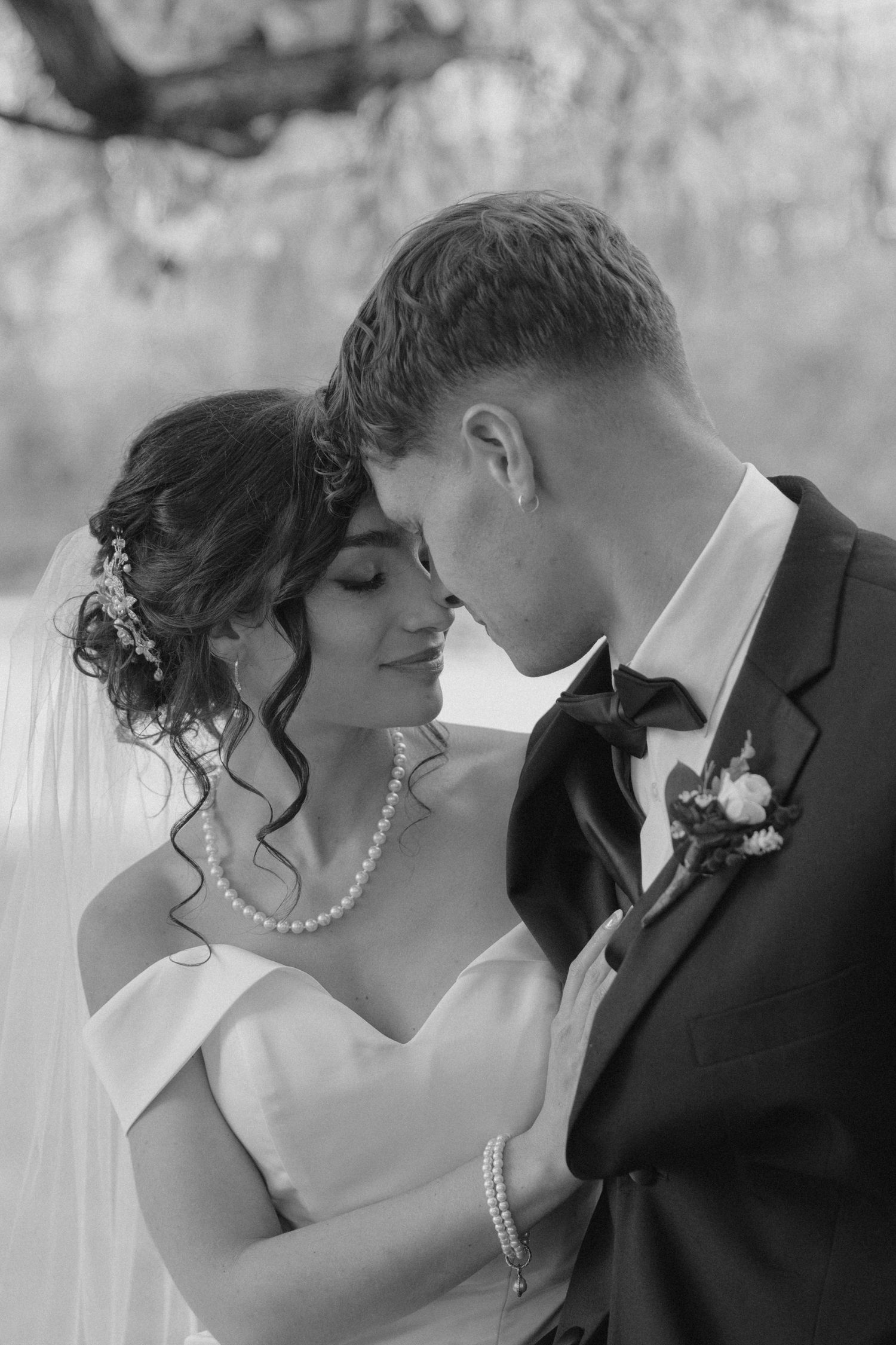 Bride in pearl necklace and groom in black tuxedo share intimate forehead touch in black and white portrait.