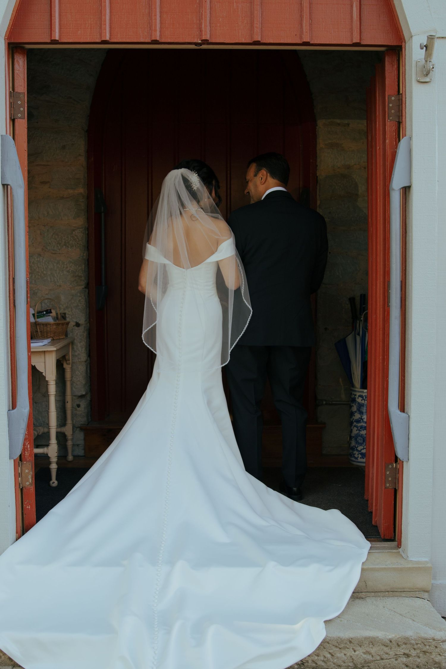 Bride in elegant mermaid gown with long veil stands with groom in church doorway after wedding ceremony.