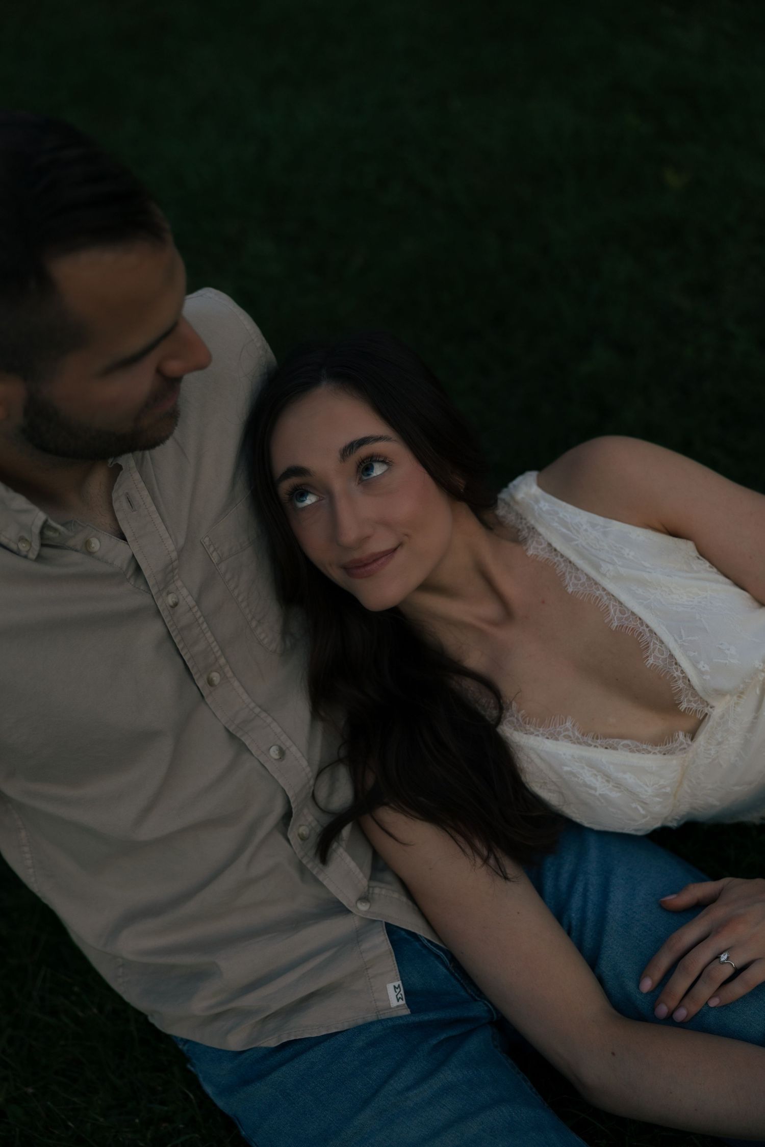 Woman in white lace dress gazes up at partner while reclining on grass at dusk.