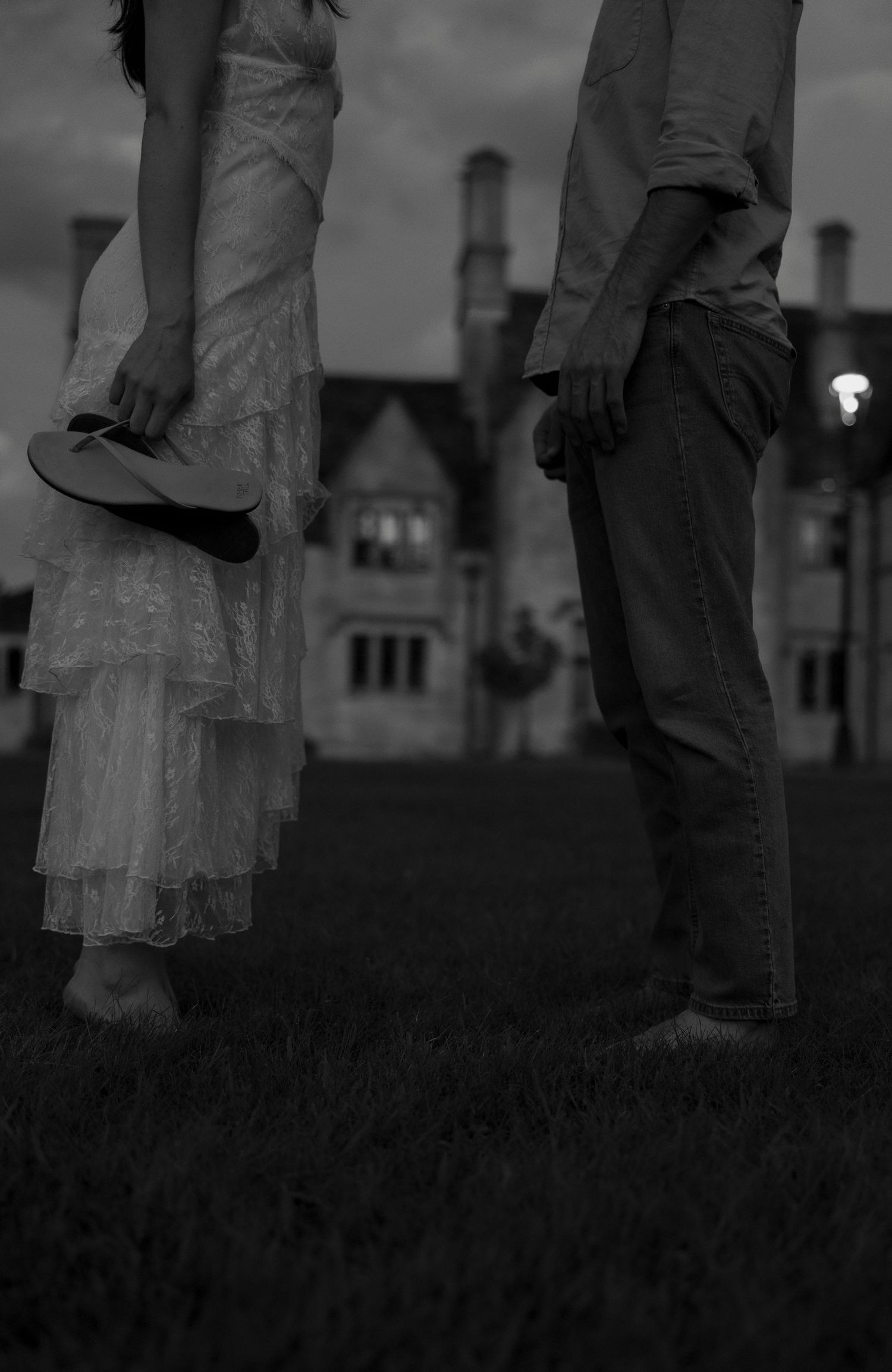 Black and white photo of a couple standing barefoot on grass holding shoes before a grand estate at twilight.