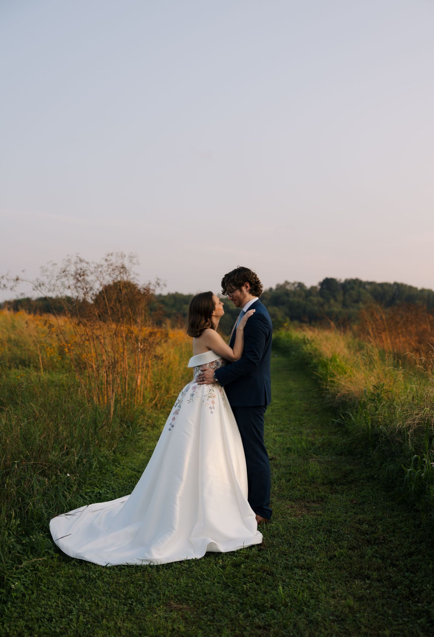 A bride and groom share a romantic embrace on a lush green path surrounded by golden meadow grasses at sunset.