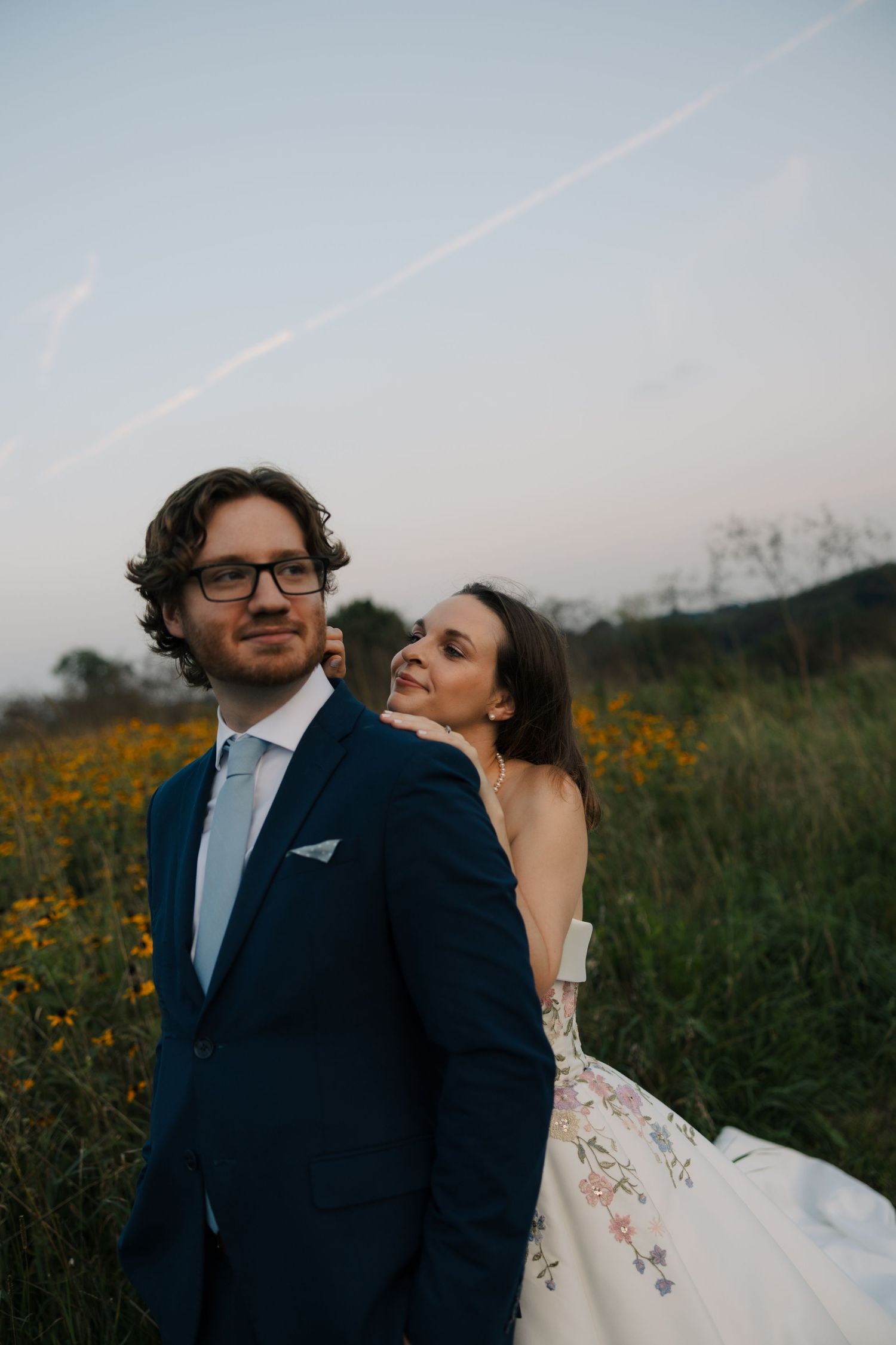 Bride in floral gown leans on groom in navy suit among wildflowers at dusk.