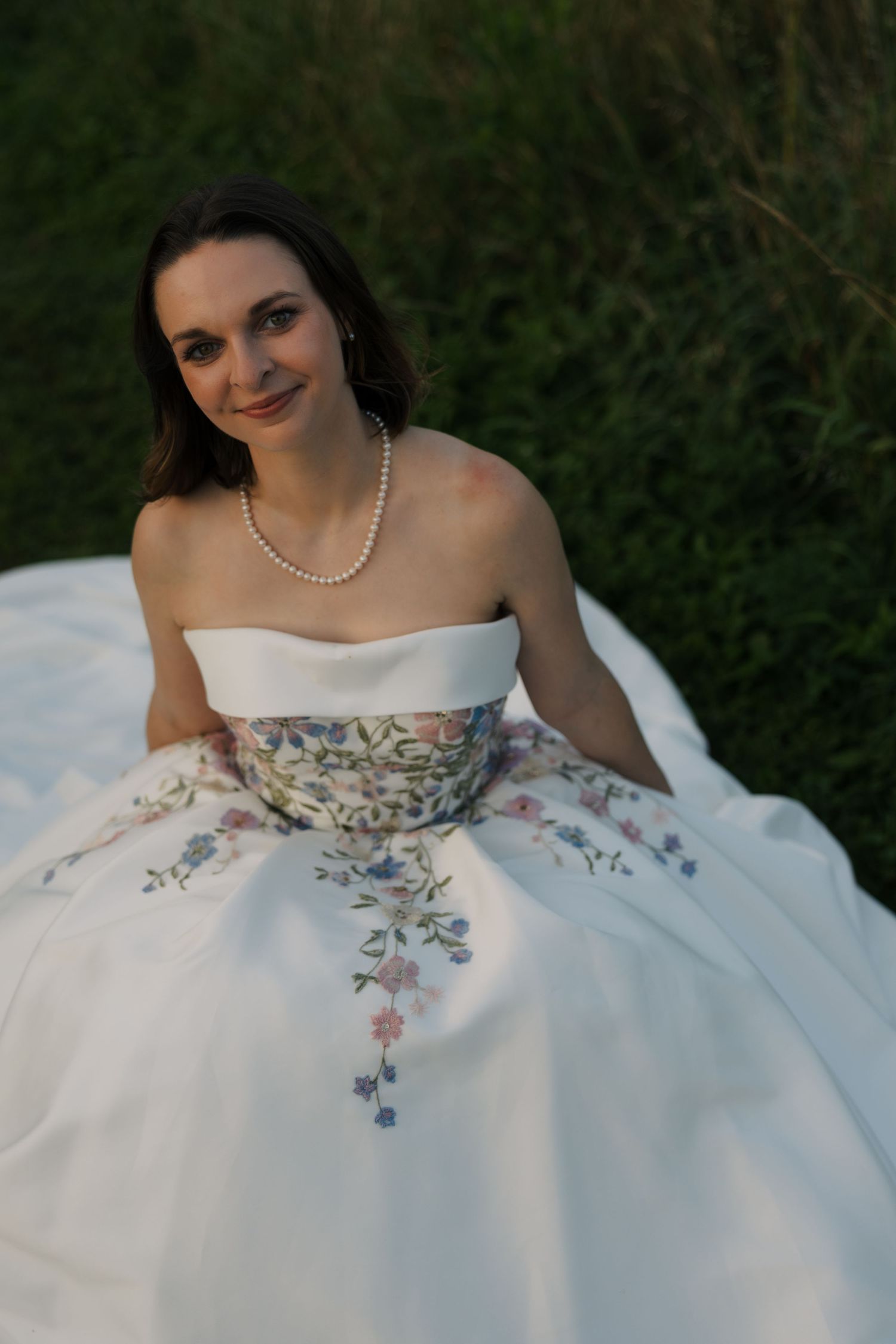 Bride sits in grass smiling, wearing strapless floral embroidered white ball gown and pearl necklace at dusk.