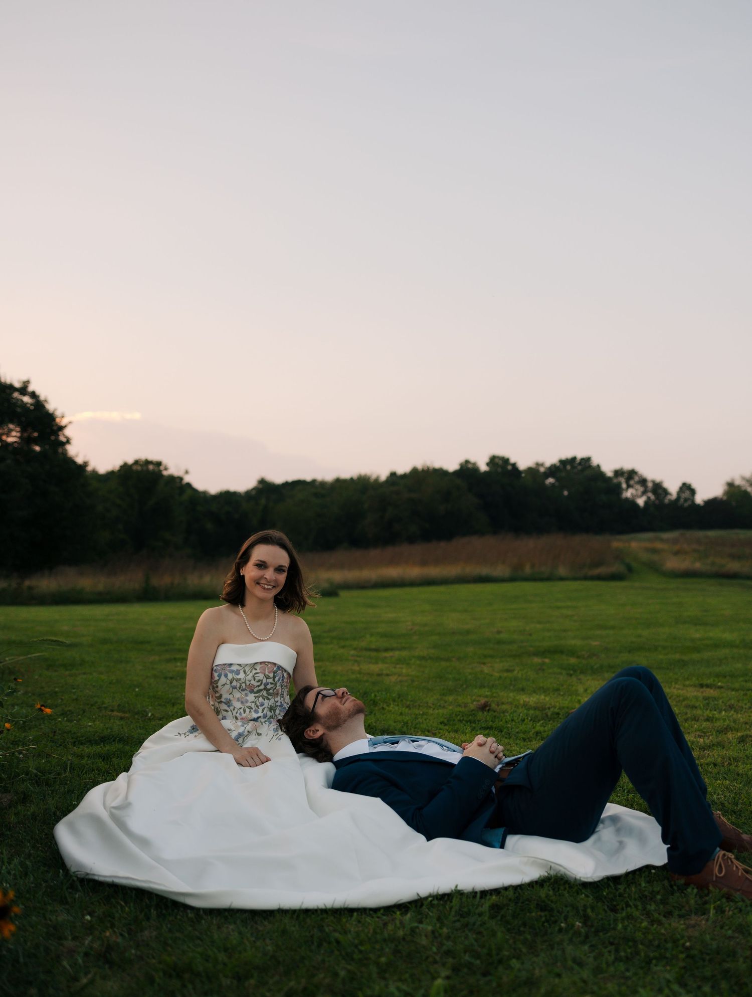 Bride in strapless floral gown sits smiling as groom lies across her lap on grass at sunset.