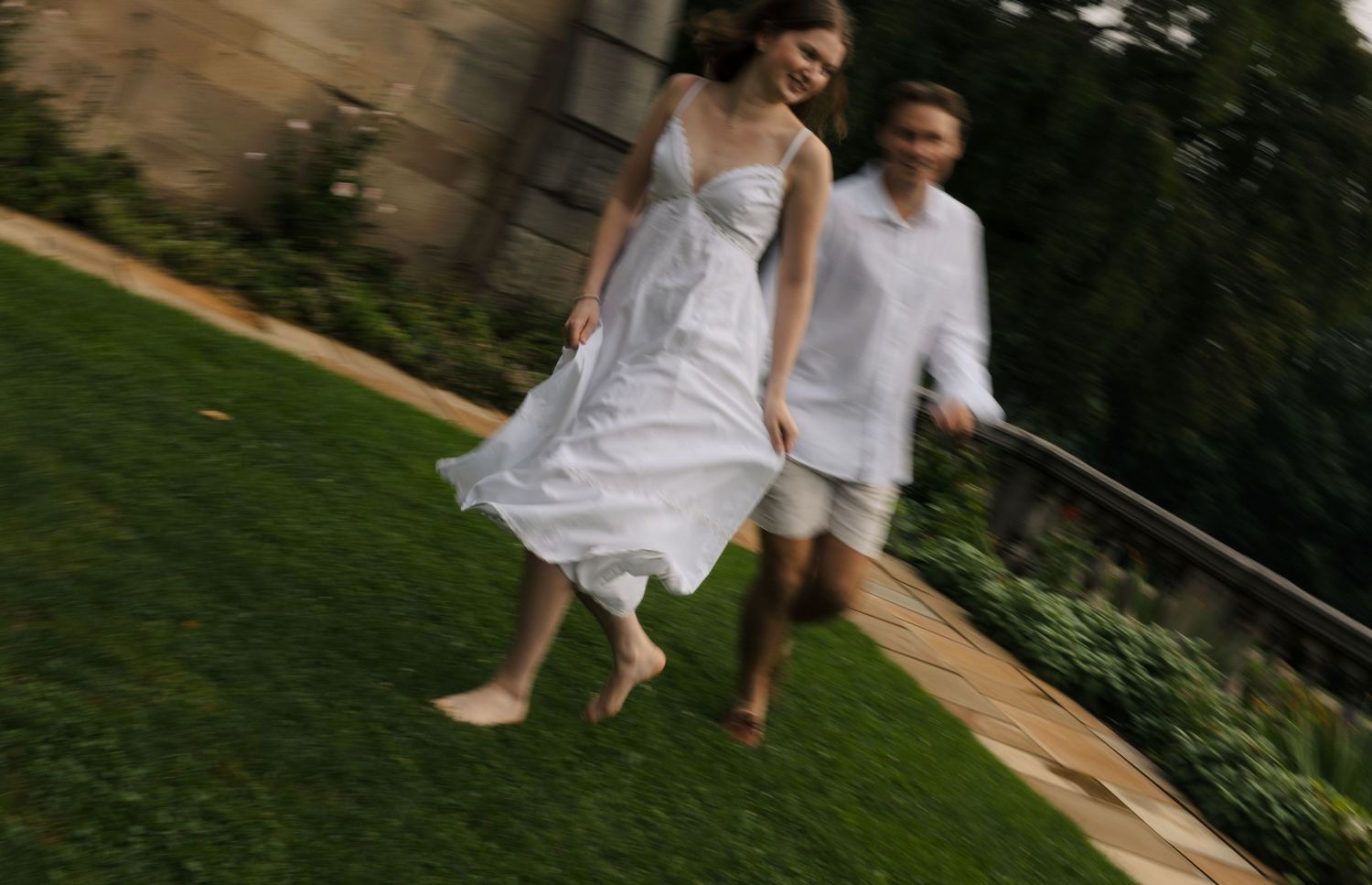 Blurred motion shot of couple running barefoot on green grass near historic stone building.