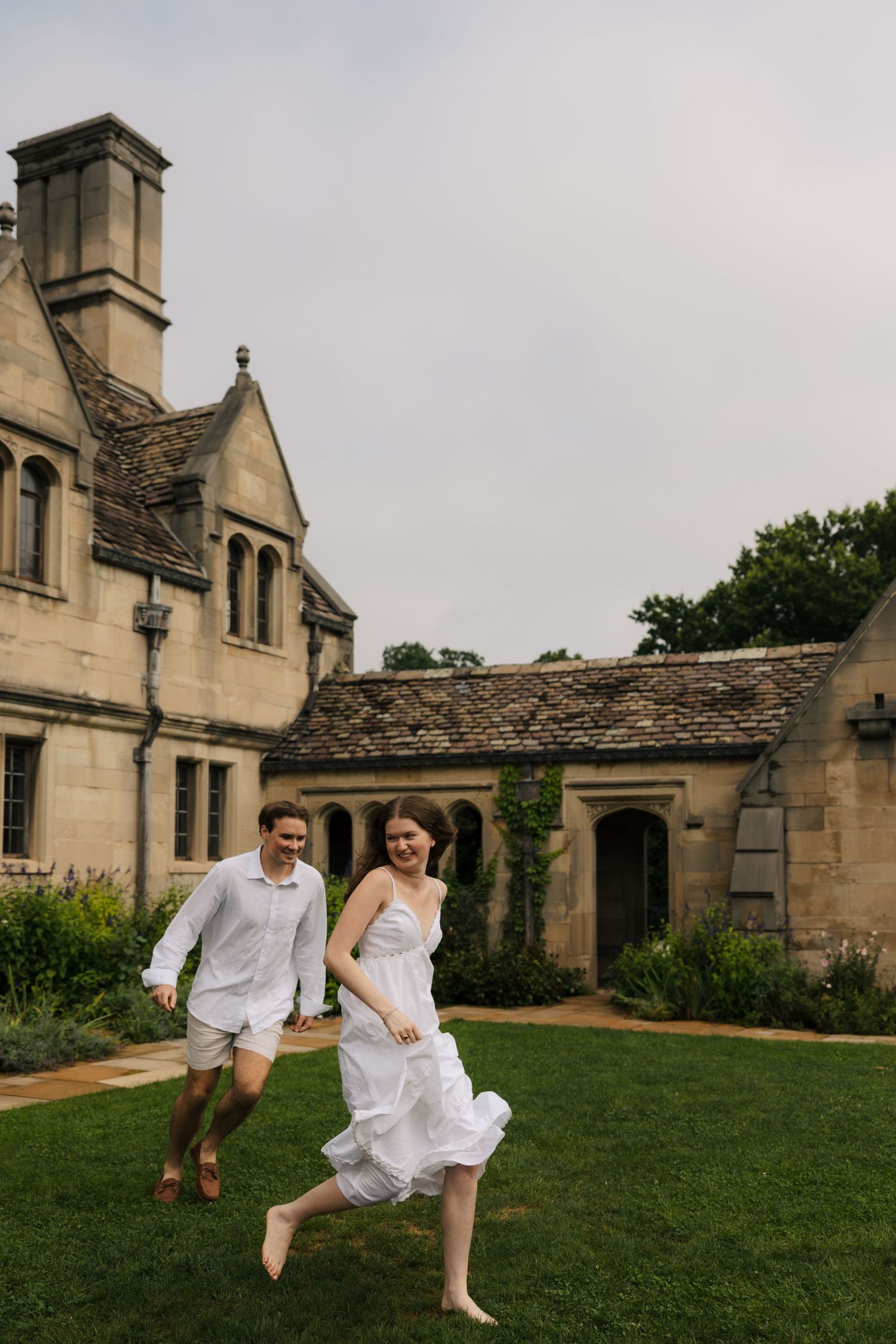 Barefoot couple in white run hand-in-hand across lawn in front of elegant stone manor house.