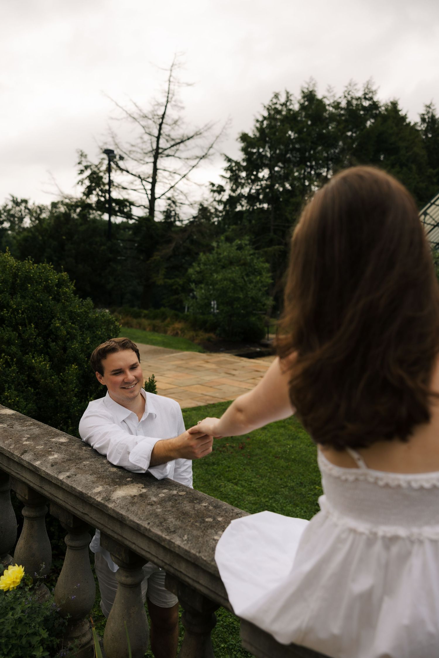 Man smiling while holding woman's hand over a stone balcony railing in a garden setting.