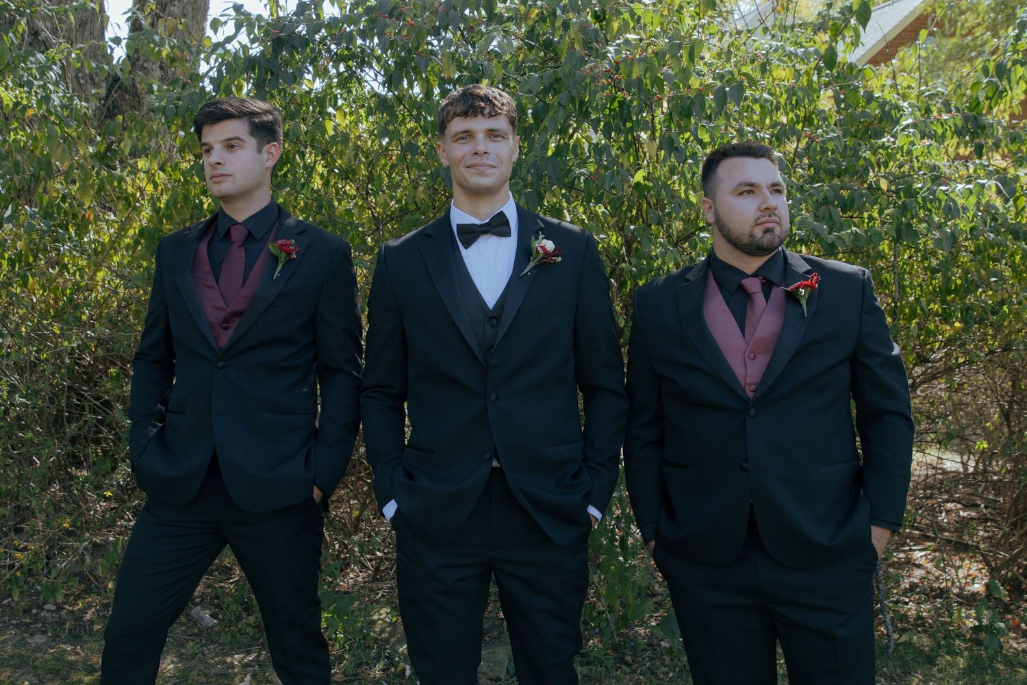 Three groomsmen in black suits and burgundy accents stand together in an outdoor garden setting.