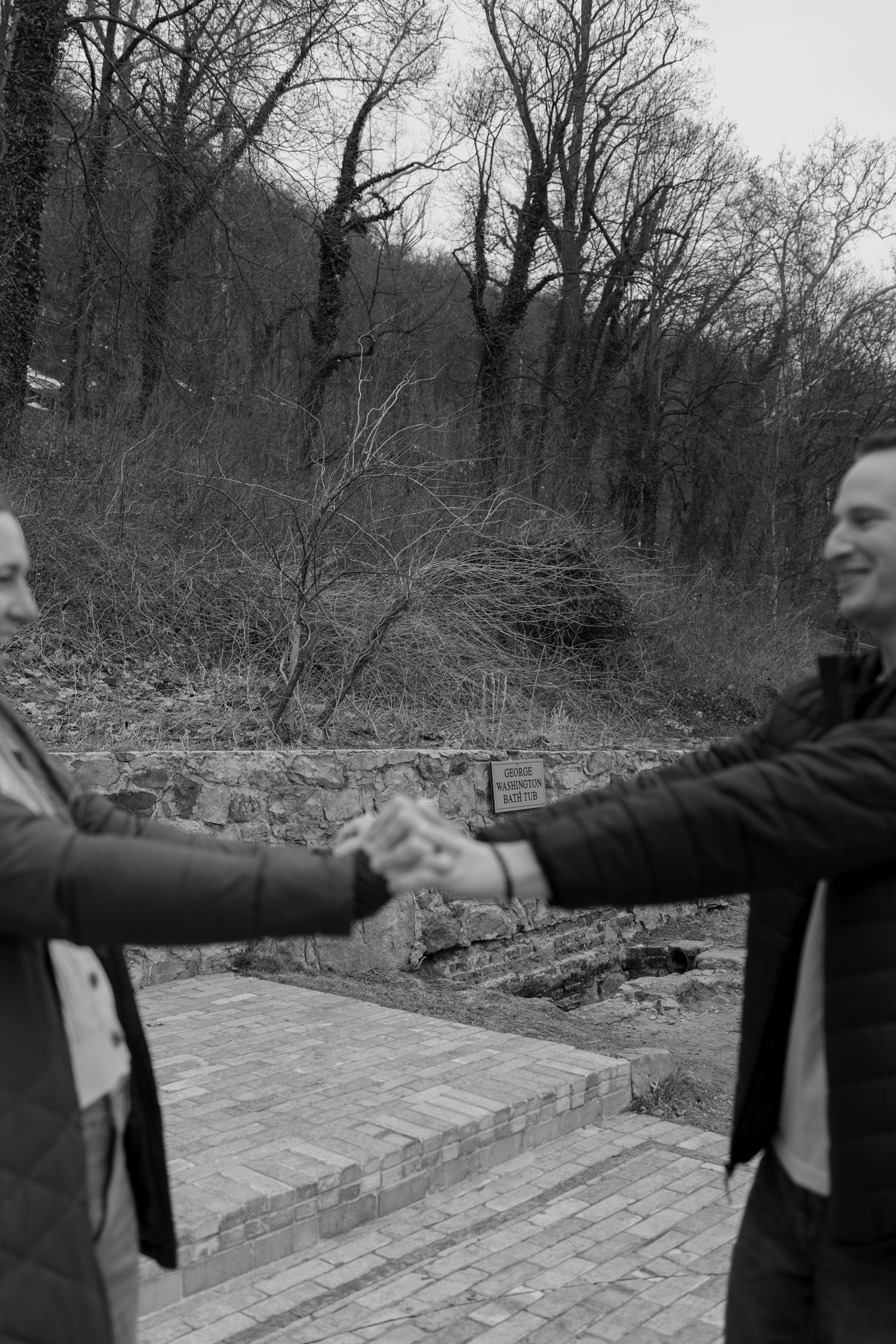 Two people in dark suits extend their arms outward while standing on a stone path in a wooded area.
