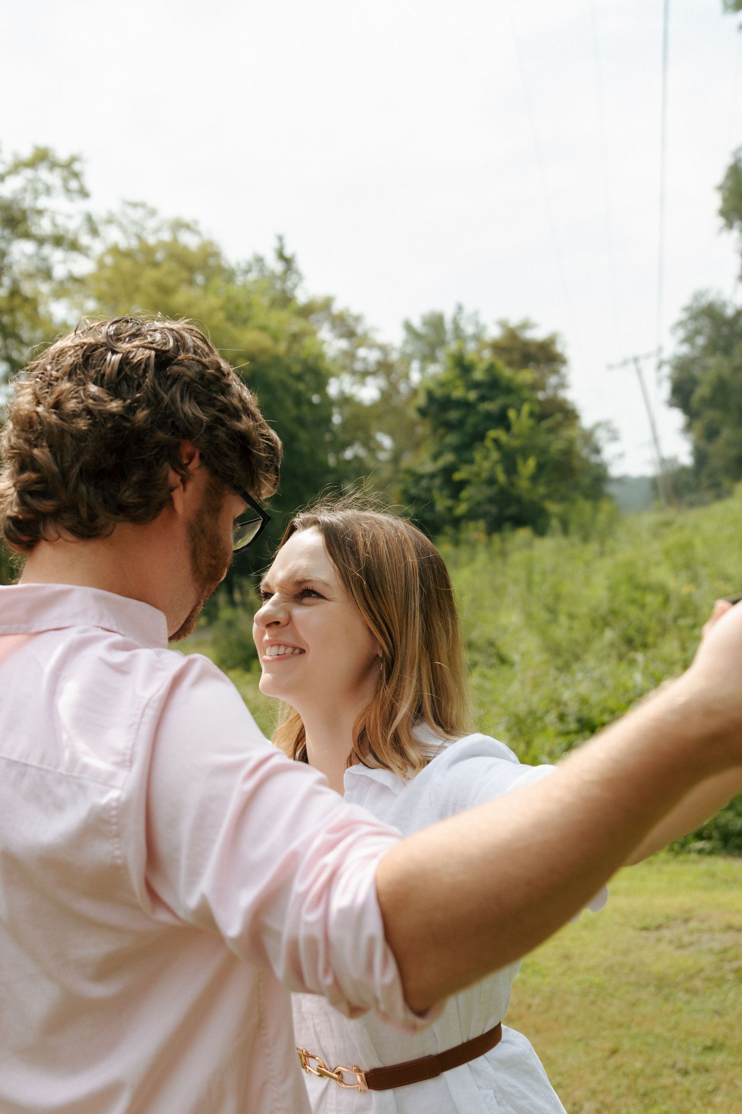 A couple in white clothing dances together joyfully in a sunny outdoor setting with green foliage in the background.