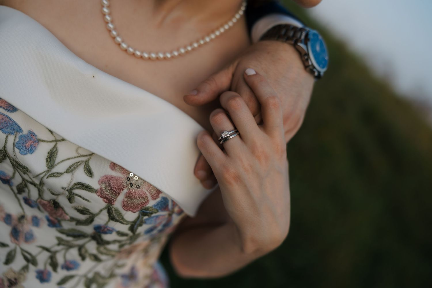 Close-up of couple holding hands showing diamond engagement ring and wedding band, pearl necklace visible.