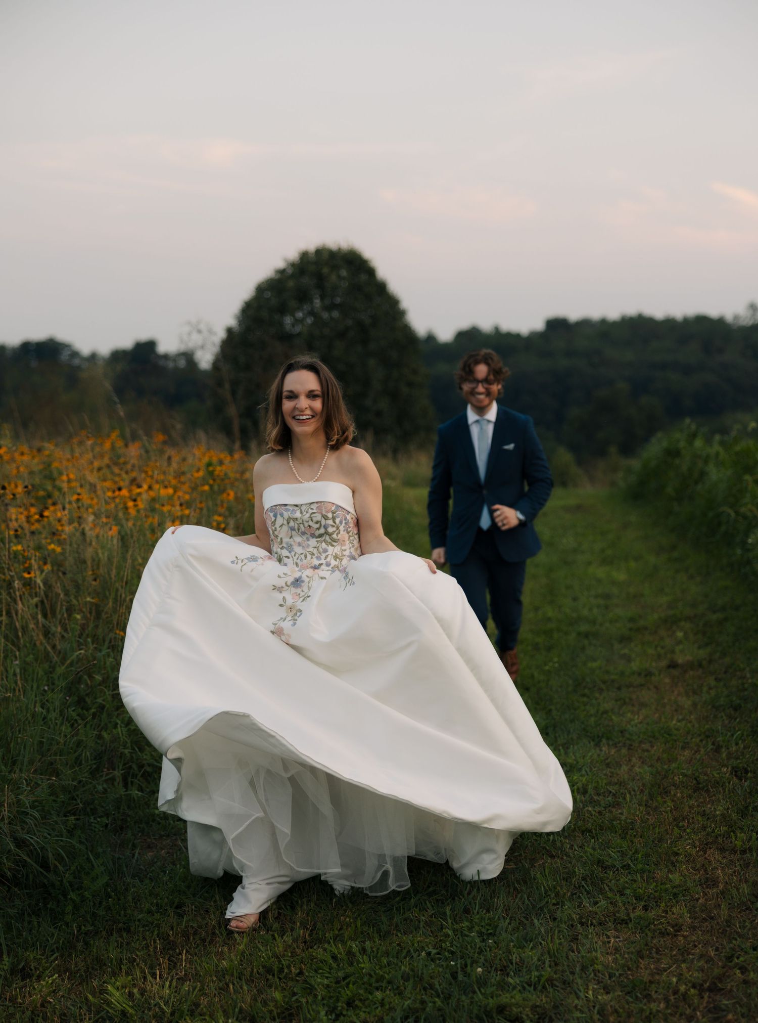 Bride in floral white gown walks through sunflower field with groom in navy suit at sunset.