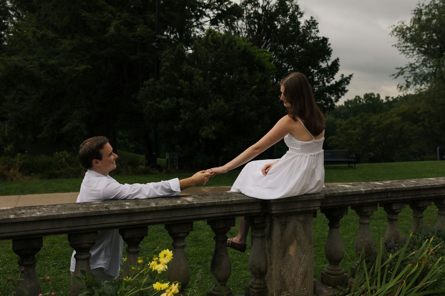 Couple holding hands across a stone balustrade in a lush garden, sharing a romantic moment.