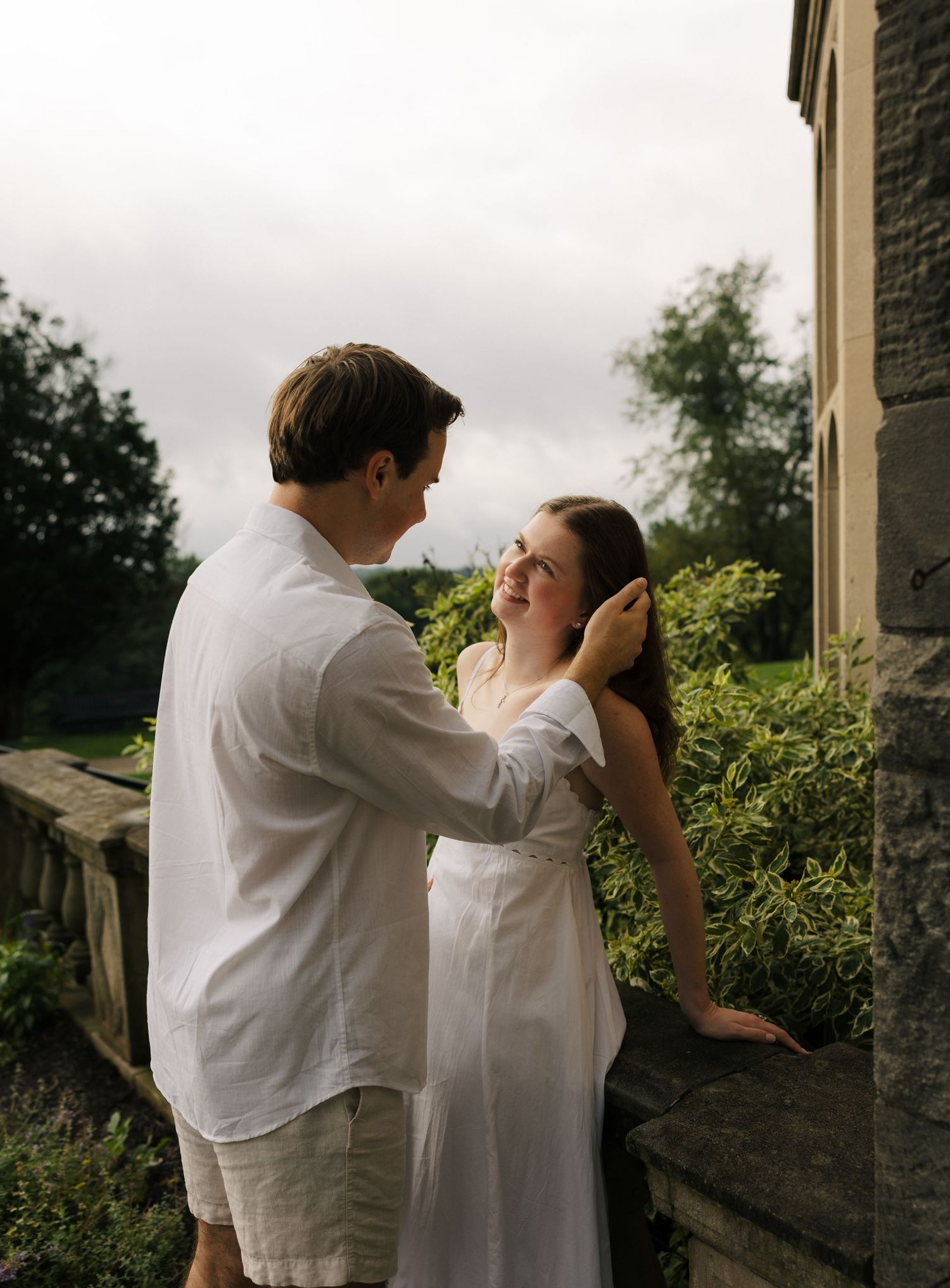 A couple in white clothing share a tender moment outdoors, the man gently touching the smiling woman's face near a stone wall.